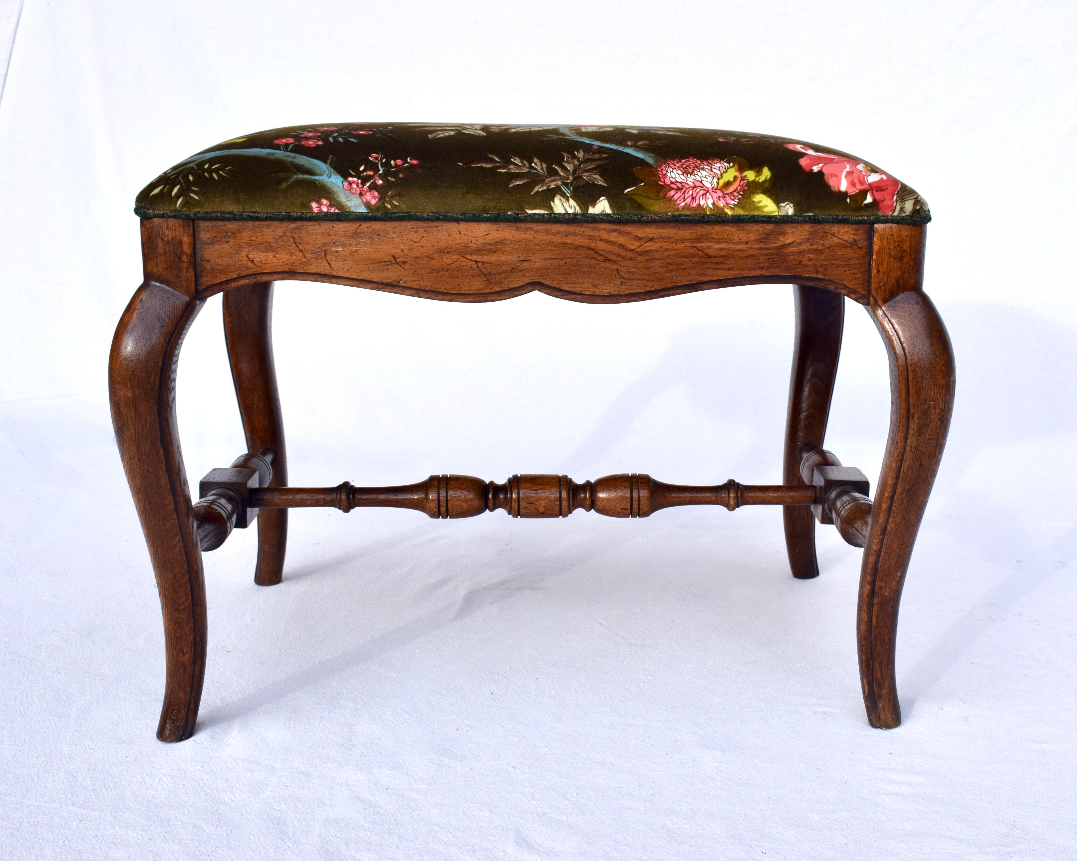 Solid Oak French style bench stool ottoman upholstered in Brunschwig & Fils floral velvet. Striking ground color is of deep olive with an array of complimenting salmon, French blue & yellows. Lovely turned H stretcher. 