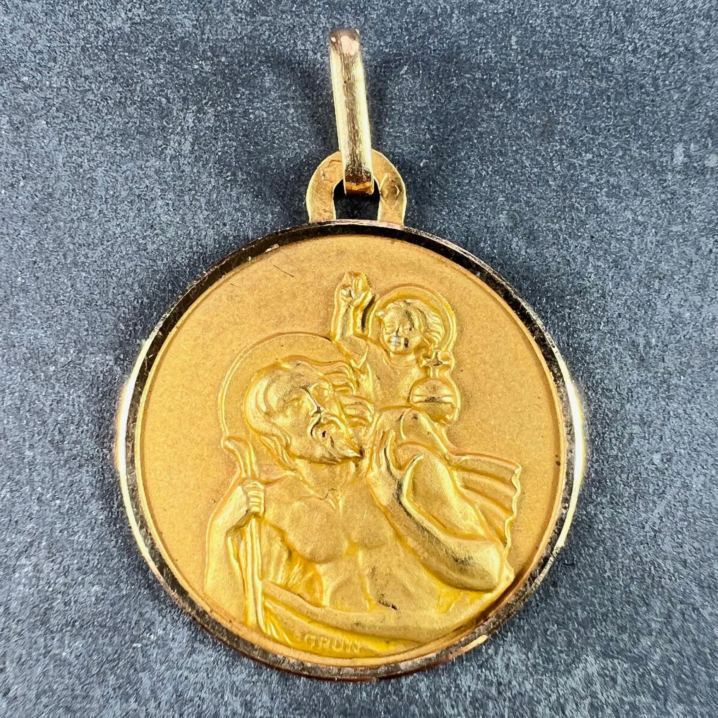 A French 18 karat (18K) yellow gold charm pendant depicting St Christopher as he carries the infant Christ across a river. Signed Grun. Stamped with the eagle’s head for French manufacture and 18 karat gold, and an unclear maker’s mark. The sculpted