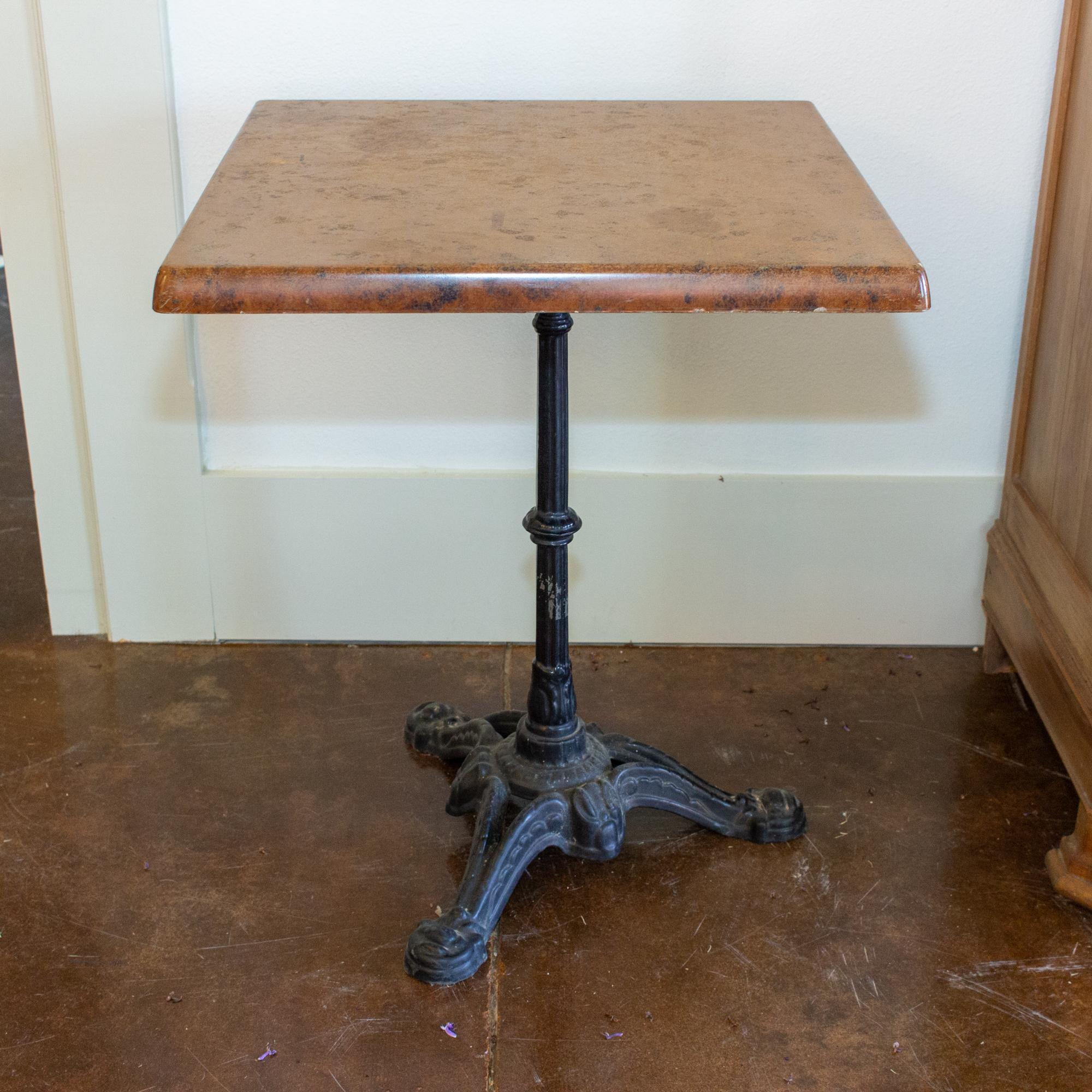 Each of these vintage French bistro tables has a black cast iron base with a square top. The tops appear to be moulded fiberglass with a faux stone design, with flared edges that mimic finished marble in a warm sienna tone. The stylized three-foot