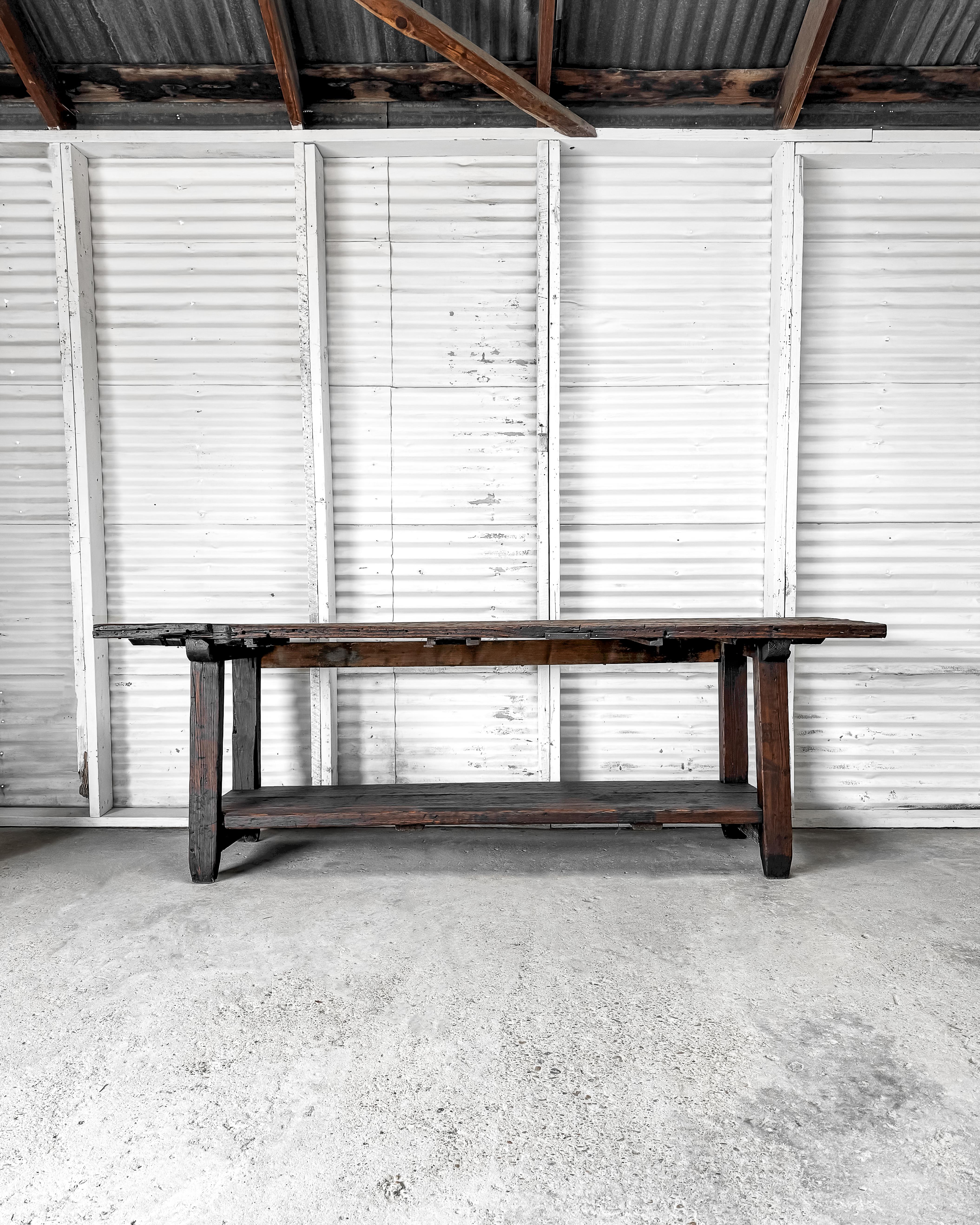 This rustic workbench, once utilized in a leather workshop in France, embodies strength and durability. Its substantial 3-board top sits atop squared legs, accompanied by a lower planked shelf that runs along the base. Bearing the marks of its rich