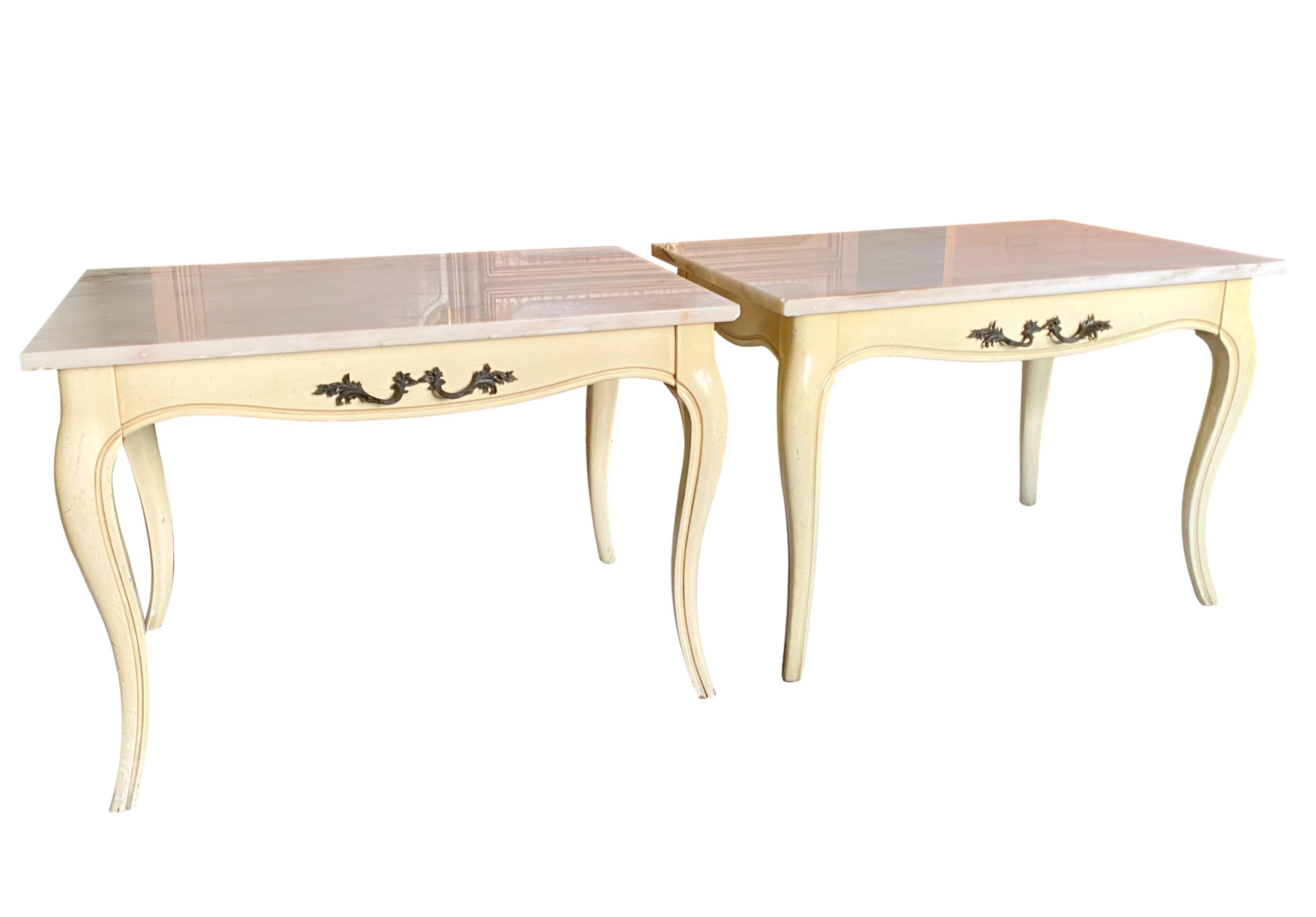A vintage pair of French Louis XV style marble top accent tables. Cream painted wood frames featuring cabriole legs, channeled edge details and front/back faux drawer handles in bronze. Marble tops are an off-white shade with veins of amber, peach