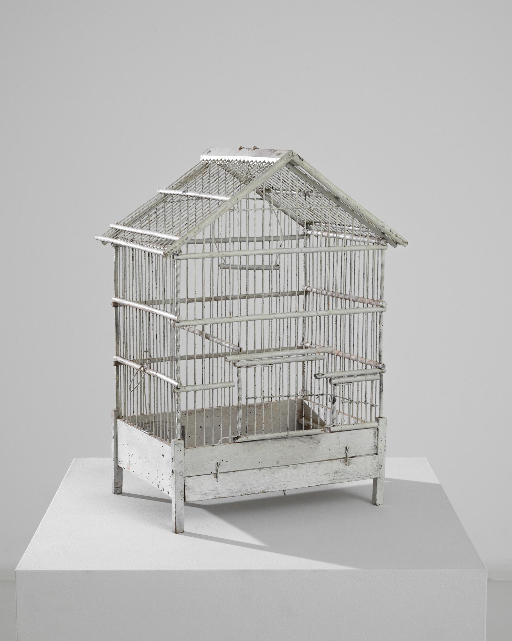 Whimsical and nostalgic, this metal birdcage brings French country charm to a space. Made at the turn of the century, the white wire silhouette suggests a cottage with a peaked roof. A variety of rods and swings would have offered the original