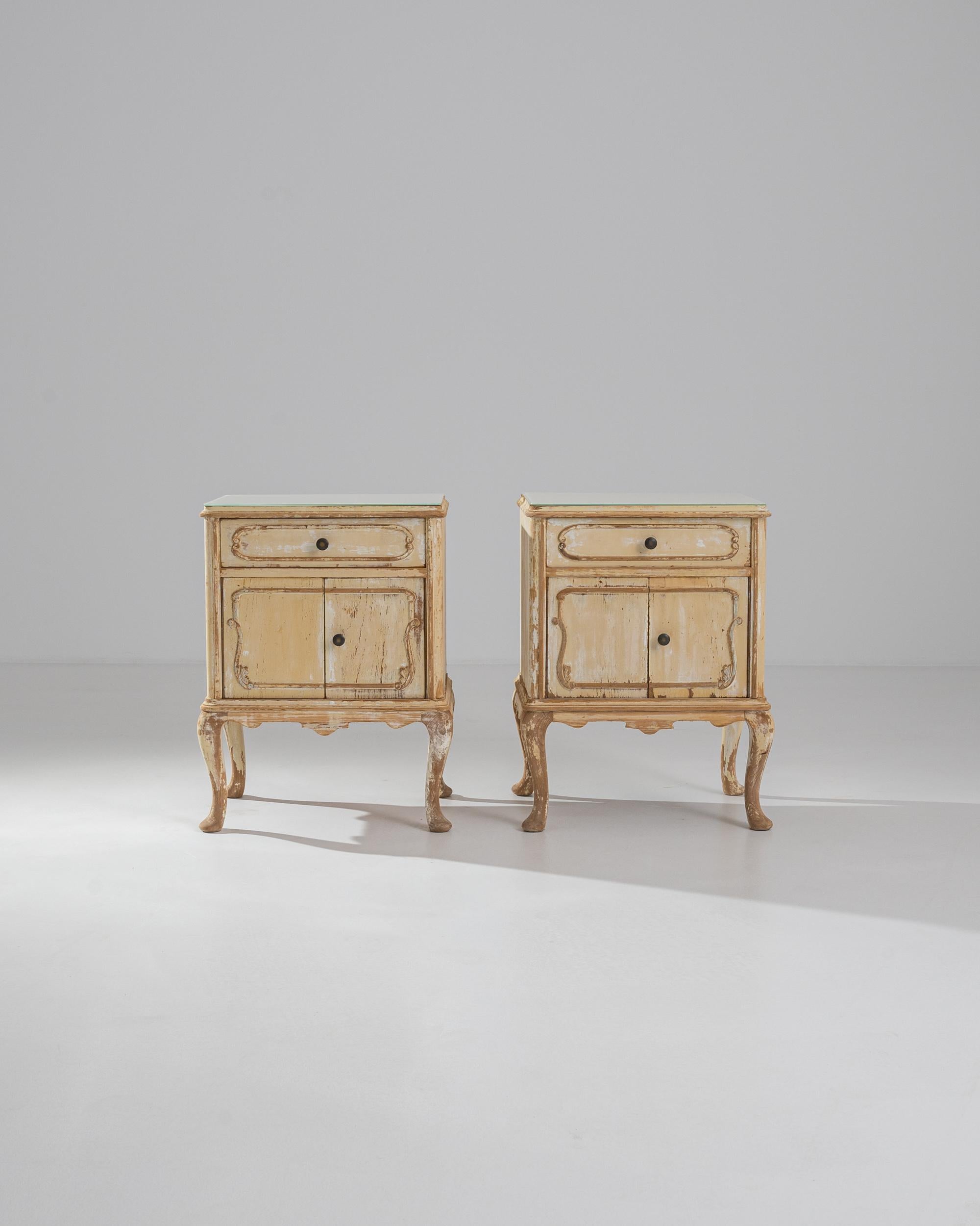 A pair of 20th century wood bedside tables with white glass tops. A creamy white patina spreads across these two bedside tables, creating a peachy brightness. With each table fitted with a drawer and storage compartment, they offer a functionality