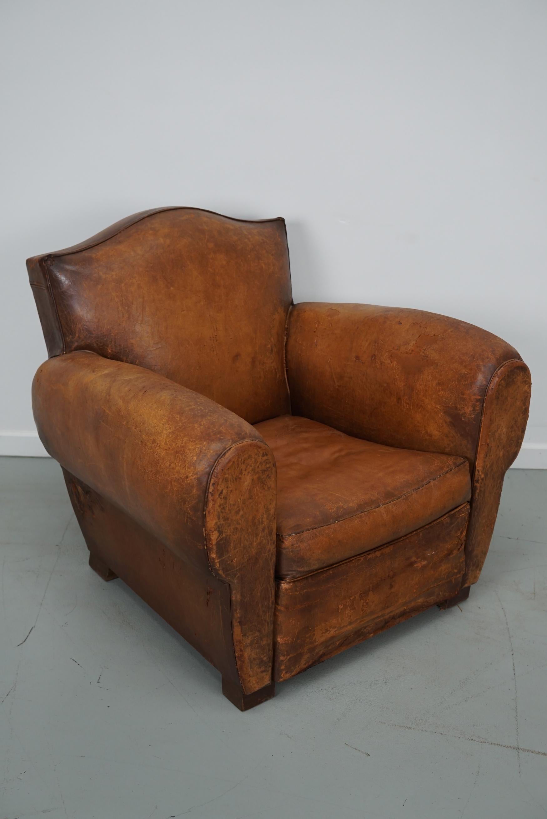 This cognac-colored leather club chair comes from France. It is upholstered with cognac-colored leather and features metal rivets and wooden legs. It is in a vintage worn condition with many signs of use, age and old repairs.