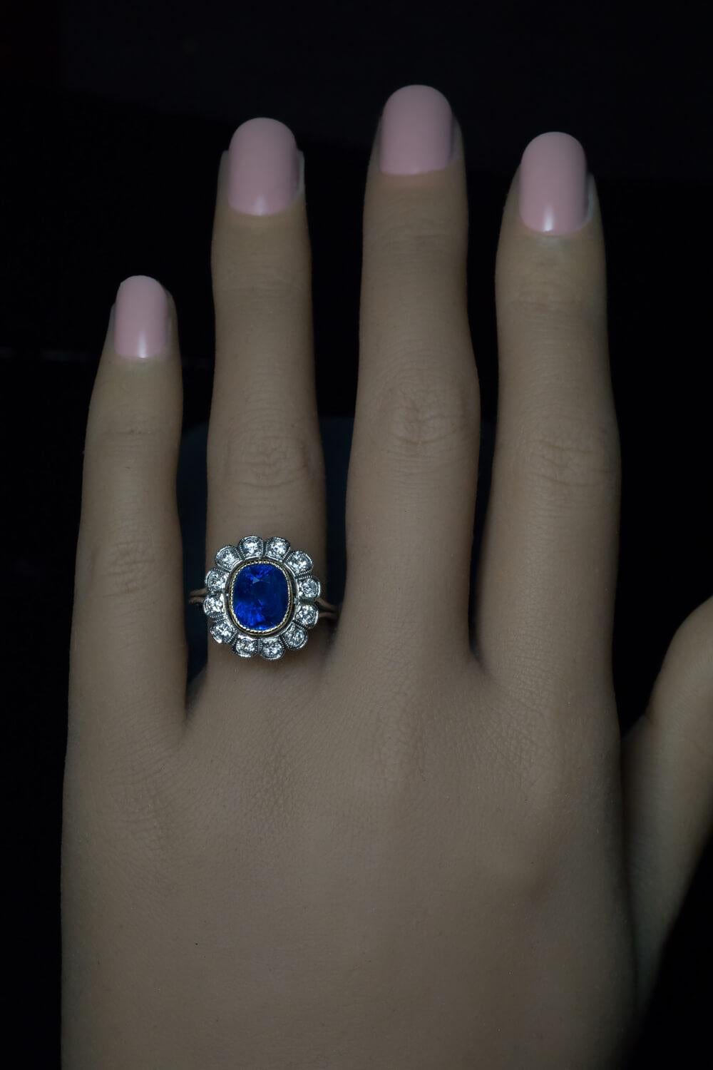France, circa 1950s.

This vintage platinum engagement ring of a classical cluster design features a cushion shaped natural unheated Ceylon sapphire of royal blue color. The sapphire is set in 18K yellow gold bezel and surrounded by bright white