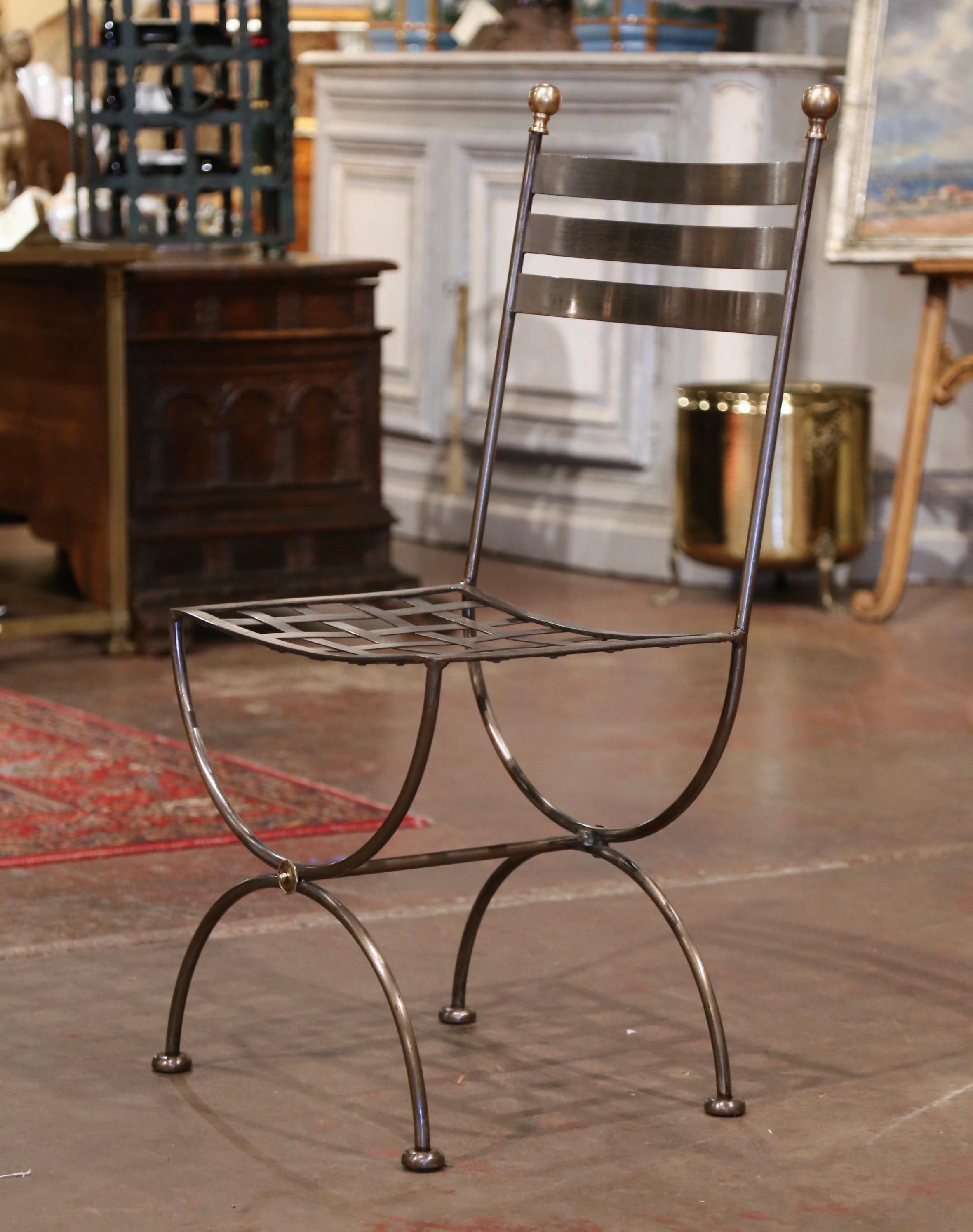 Place this elegant chair as a vanity seating, or outside on a patio! Crafted in France circa 1980 and made of wrought iron, the chair stands on forged Dagobert (or curule) legs and features three curved ladders on the back embellished with brass