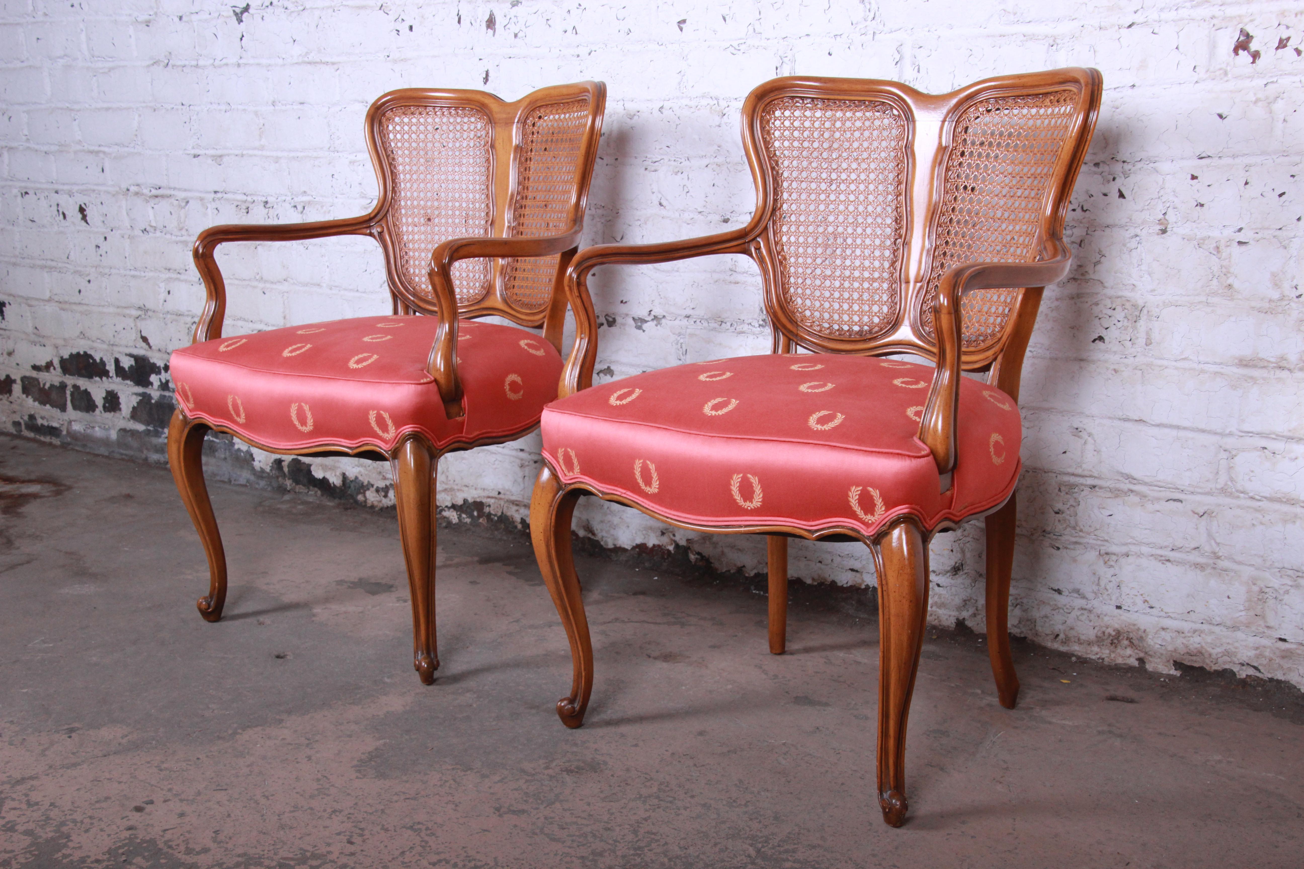 A gorgeous pair of French Provincial Louis XV style armchairs. The chairs feature solid walnut frames with a nice fanned cane back and original soft red/pink upholstery. Similar in quality and craftsmanship to Baker Furniture, although the chairs
