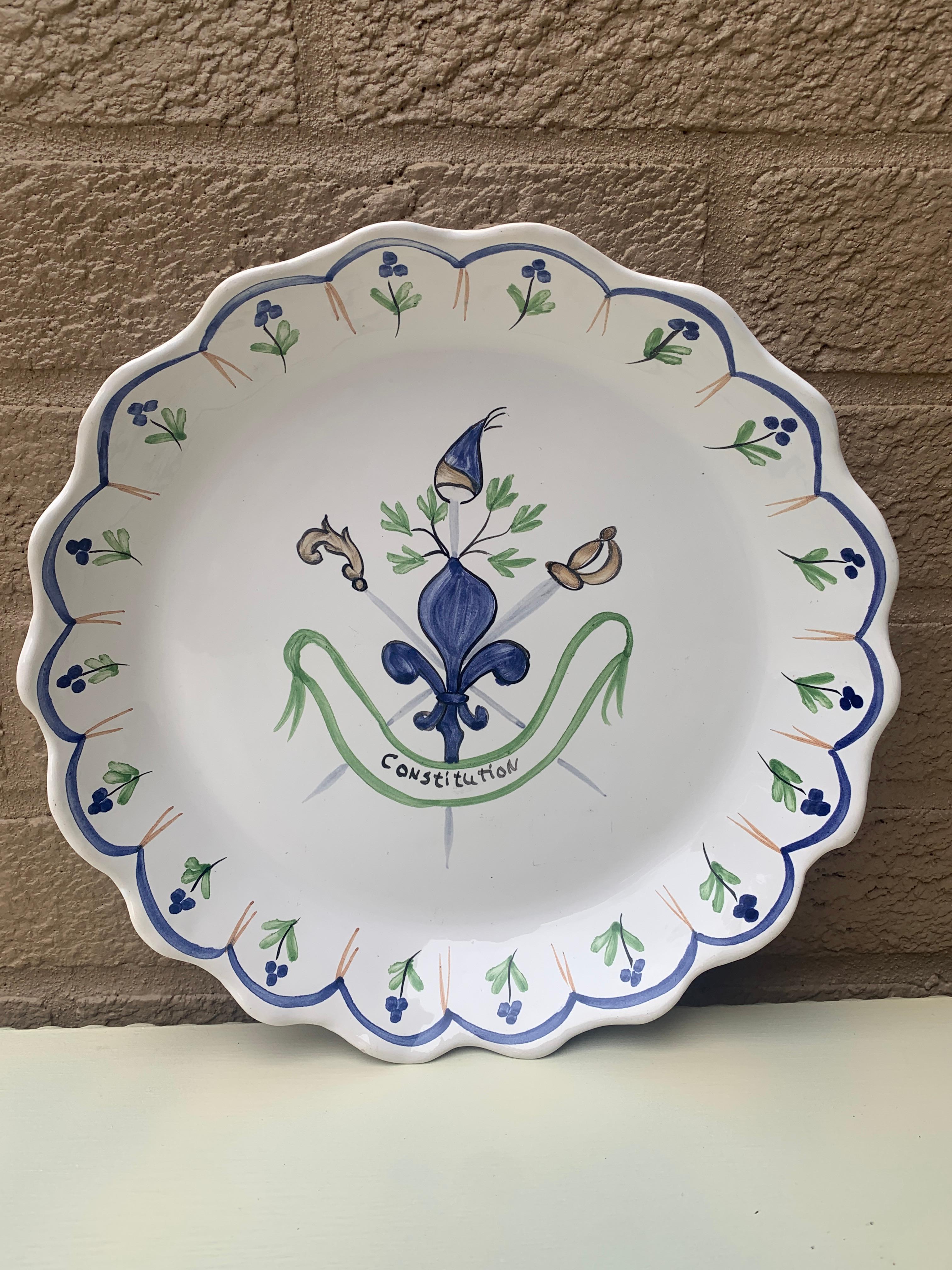 A beautiful hand painted blue, white, and green faience pottery wall plate featuring a fleur de lis, crossed swords, a French hat, and a banner that reads 