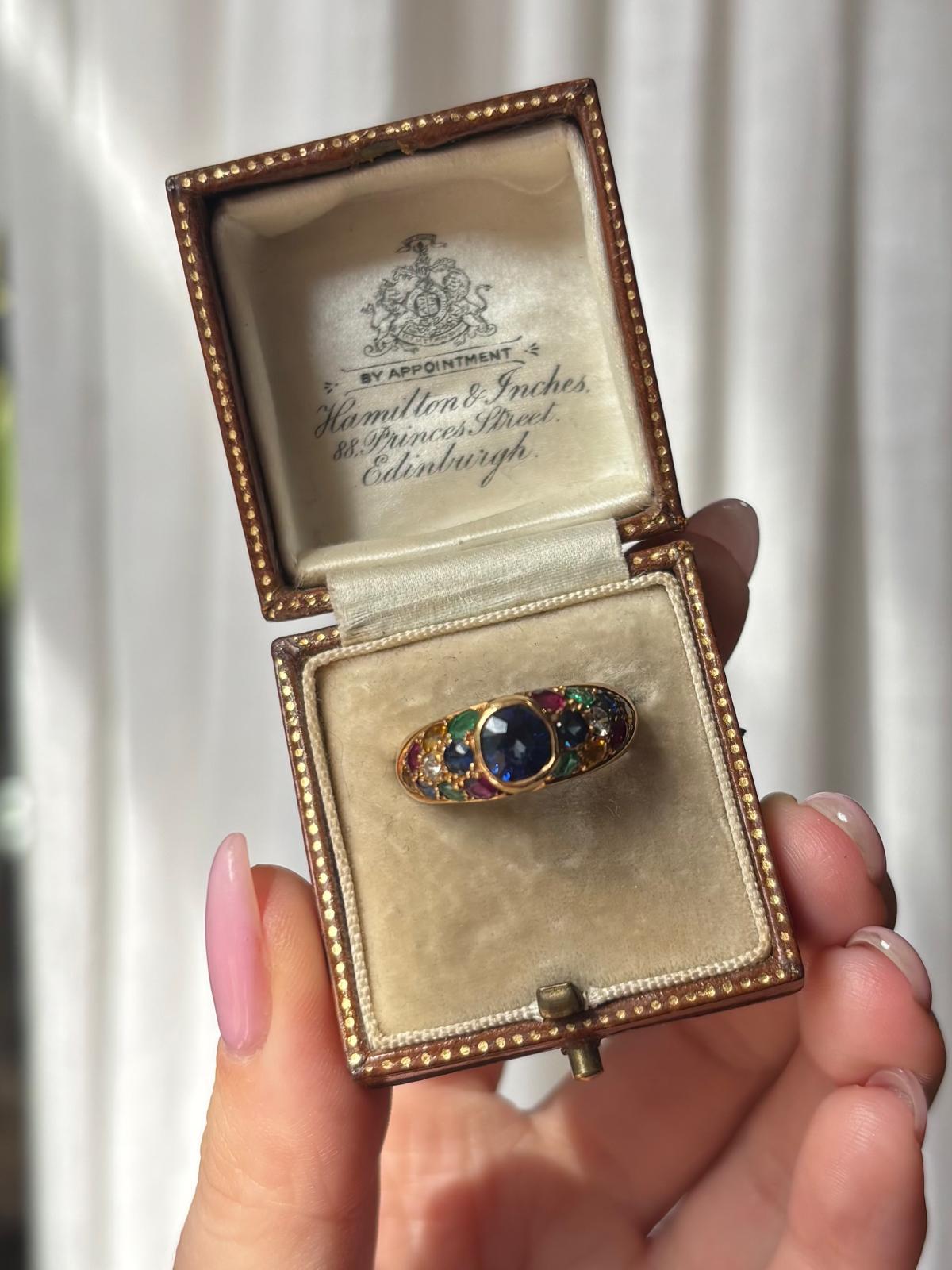 A vibrant vintage sapphire, emerald, ruby and diamond bombe ring set in 18k yellow gold. 

The ring is centrally rub over set with a sparkly royal blue coloured cushion cut sapphire. The sapphire is set in a raised bombe setting with multi gem set