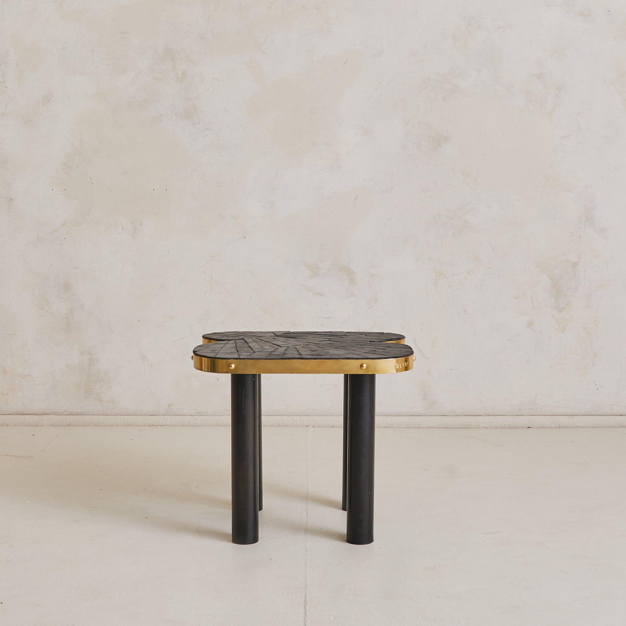 A sculptural 1990s French side table defined by bold material contrast and graphic rhythm. The clover-shaped top is composed of hand-cut slate, laid in a radiating pattern and encircled by a thick brass band. Four cylindrical legs in blackened steel