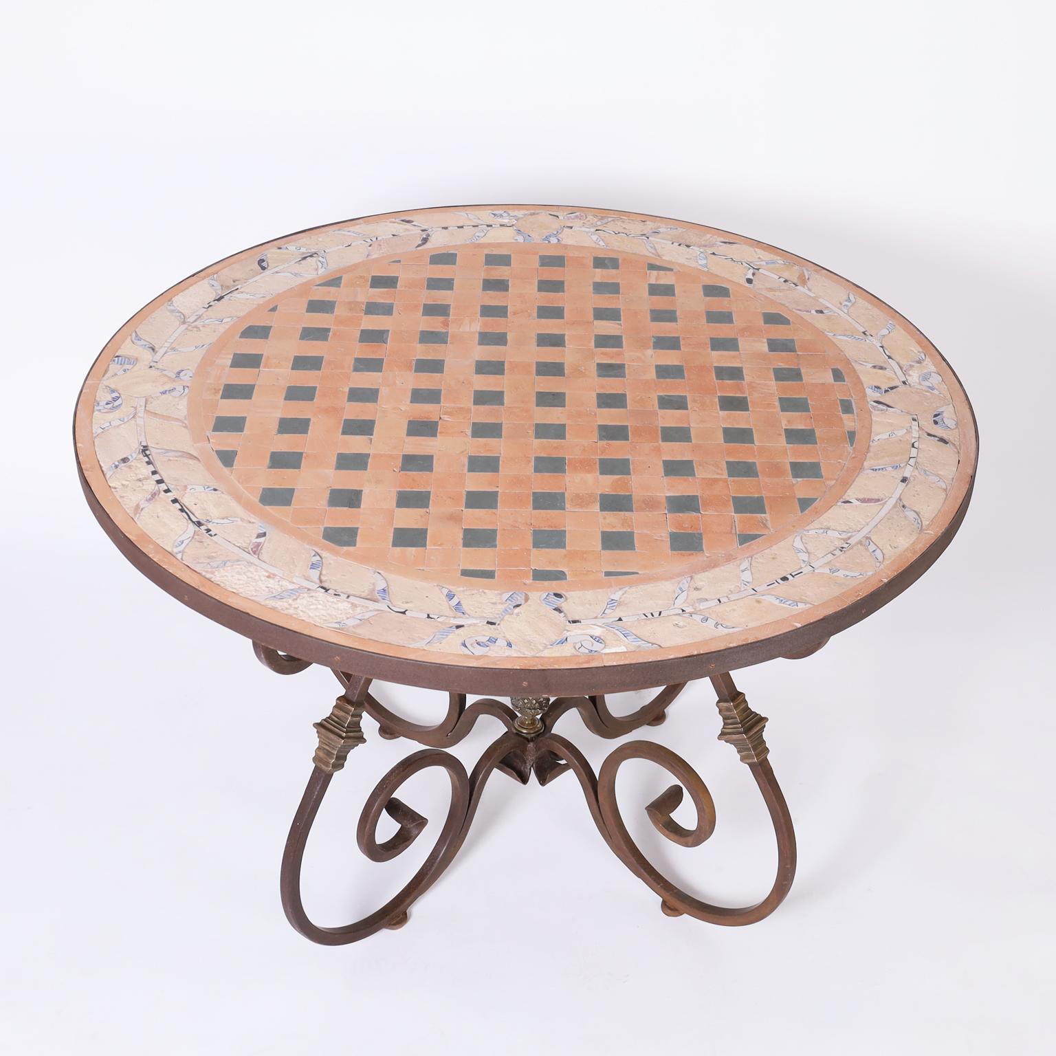 French garden table with a round top having a geometric glazed terra cotta center bordered by pottery shards in floral designs. The wrought iron base has graceful lines highlighted with bronze cuffs and pineapple at the center.