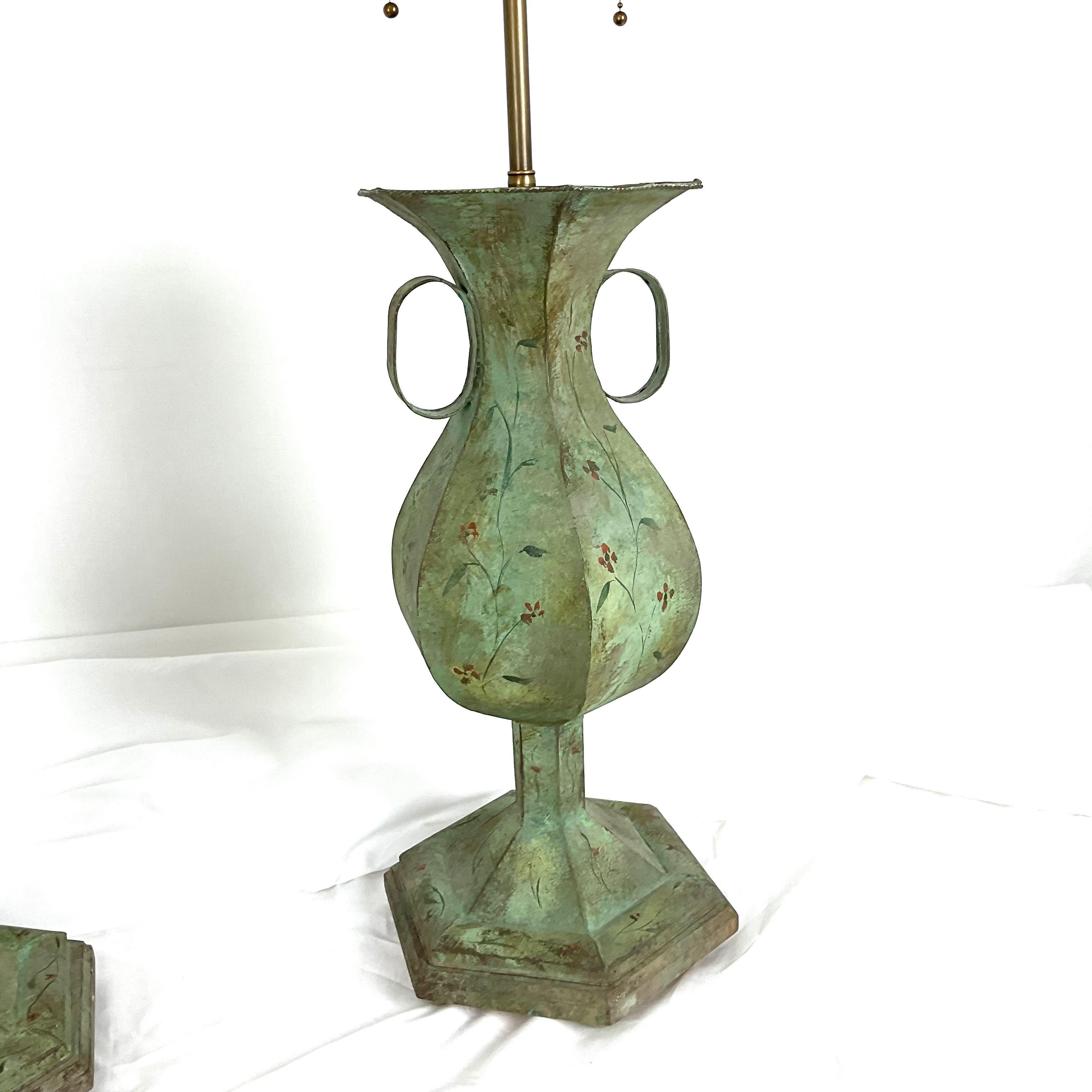 Cette étonnante paire de lampes de table tole vintage présente une décoration florale peinte à la main sur une finition patinée vert tendre. Les élégants corps en forme de balustre reposent sur des socles étagés, accentués par des poignées latérales