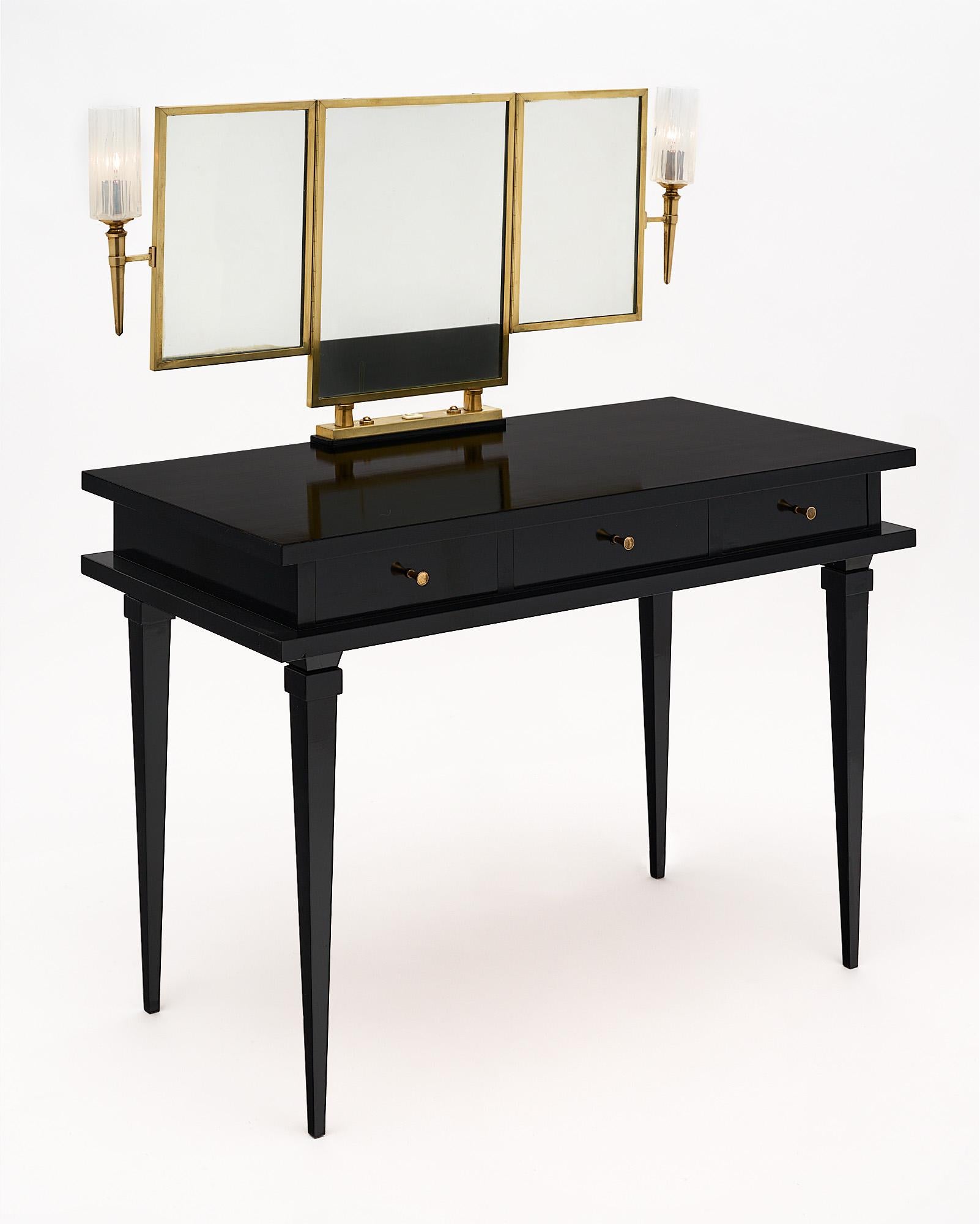 Vanity, French, made of ebonized sycamore. The vanity is in the manner of Jacques Adnet and features three dovetailed drawers and tapered legs. This elegant piece is finished in a lustrous French polish and enhanced with a triptych brass framed