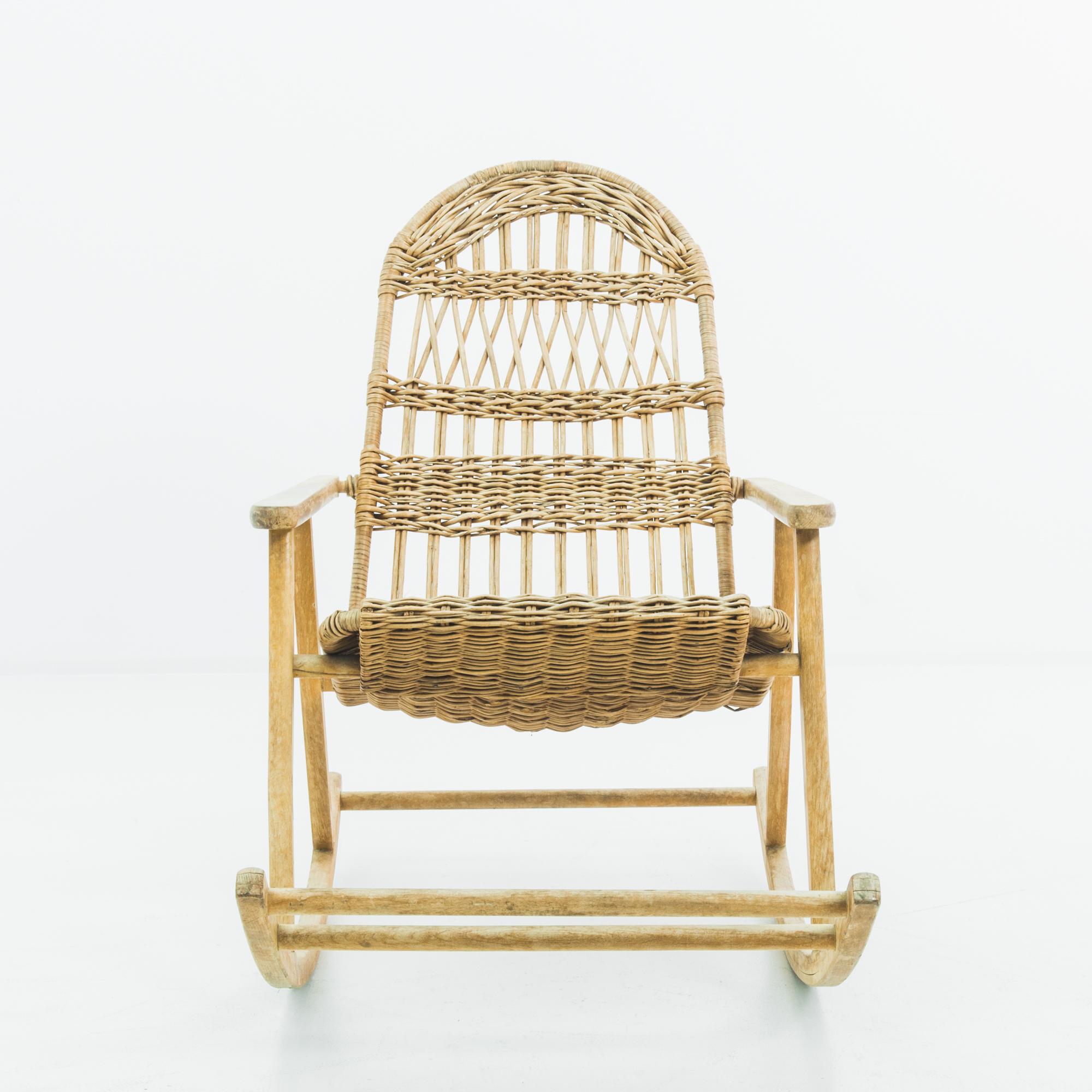 A wooden rocking armchair from France, produced circa 1960. A wicker seat woven around a wooden frame resting on a pair of elongated rockers with built-in footrest. A comfortable rocking chair built for hours of lounging in the sun or nestled by the