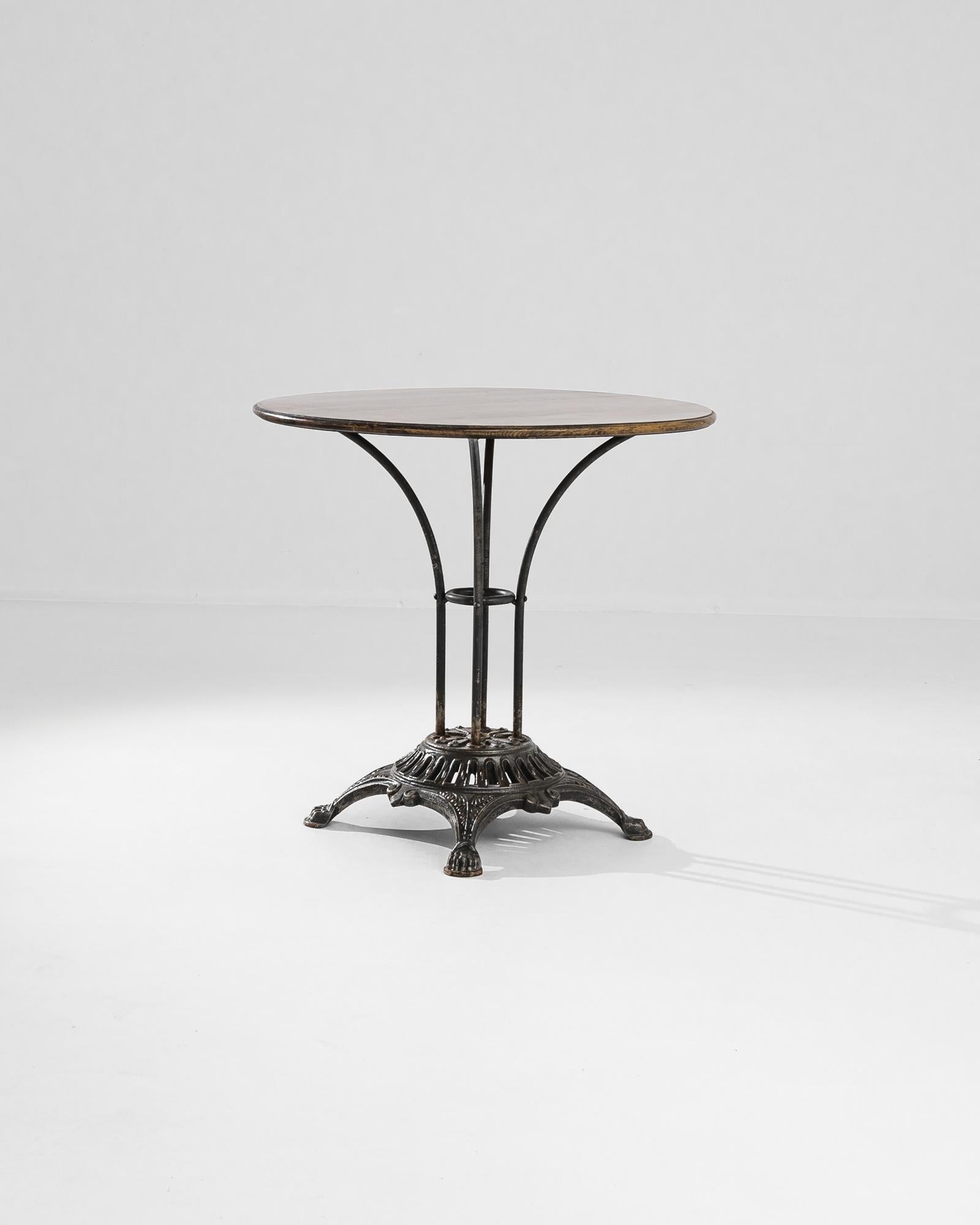 An elegant circular table from 1950s France. The polished wooden tabletop is elevated upon an arrangement of arched metal rods, set atop an elaborate cast-iron base — a bistro Silhouette with a distinguished twist. Paw feet gift a light-footed