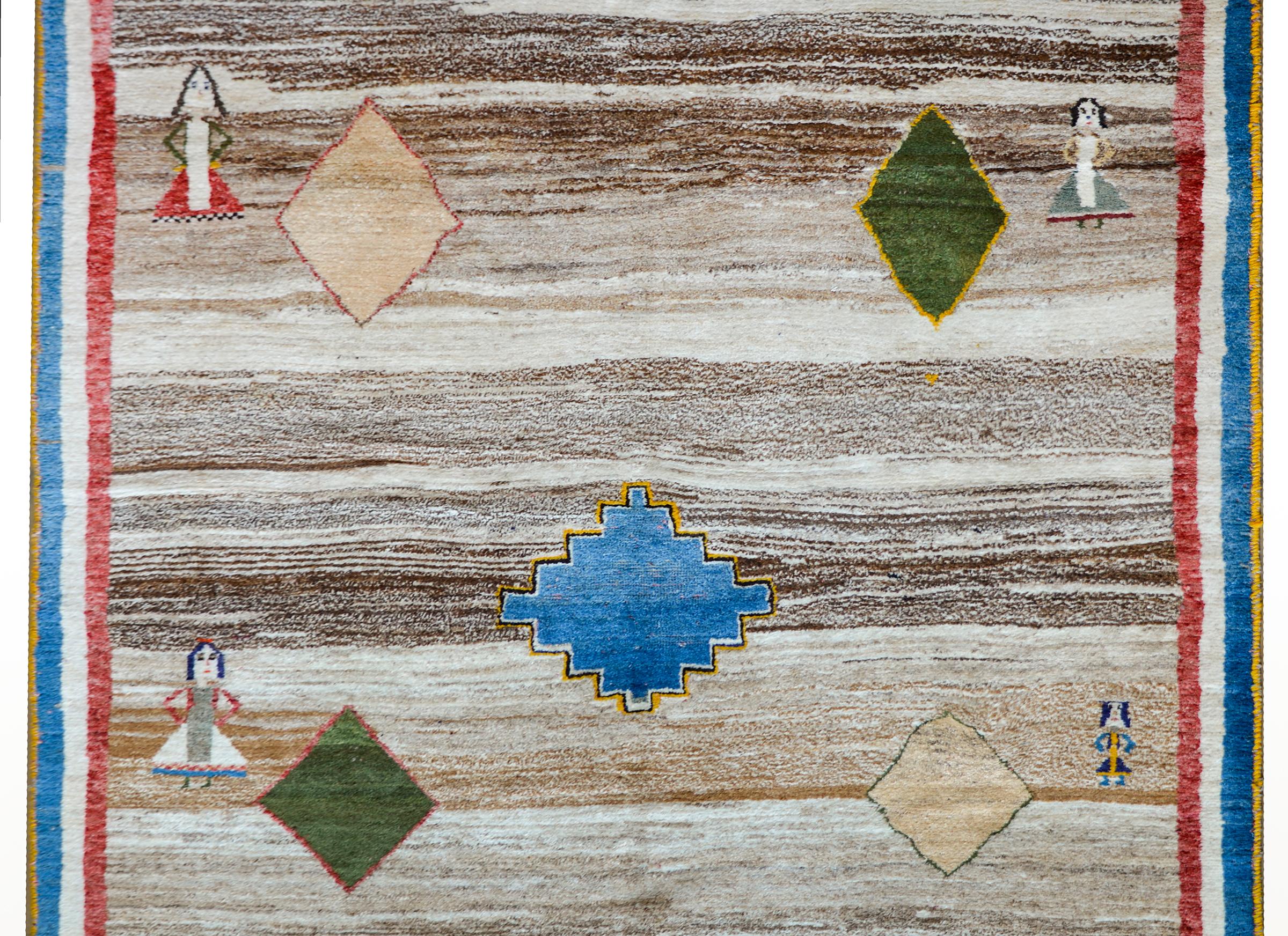 A whimsical vintage Persian Gabbeh rug with an abrash natural brown and cream background with several indigo, green, and cream diamond medallions, and four women in each corner, and surrounded by a red, white, and blue striped border.
