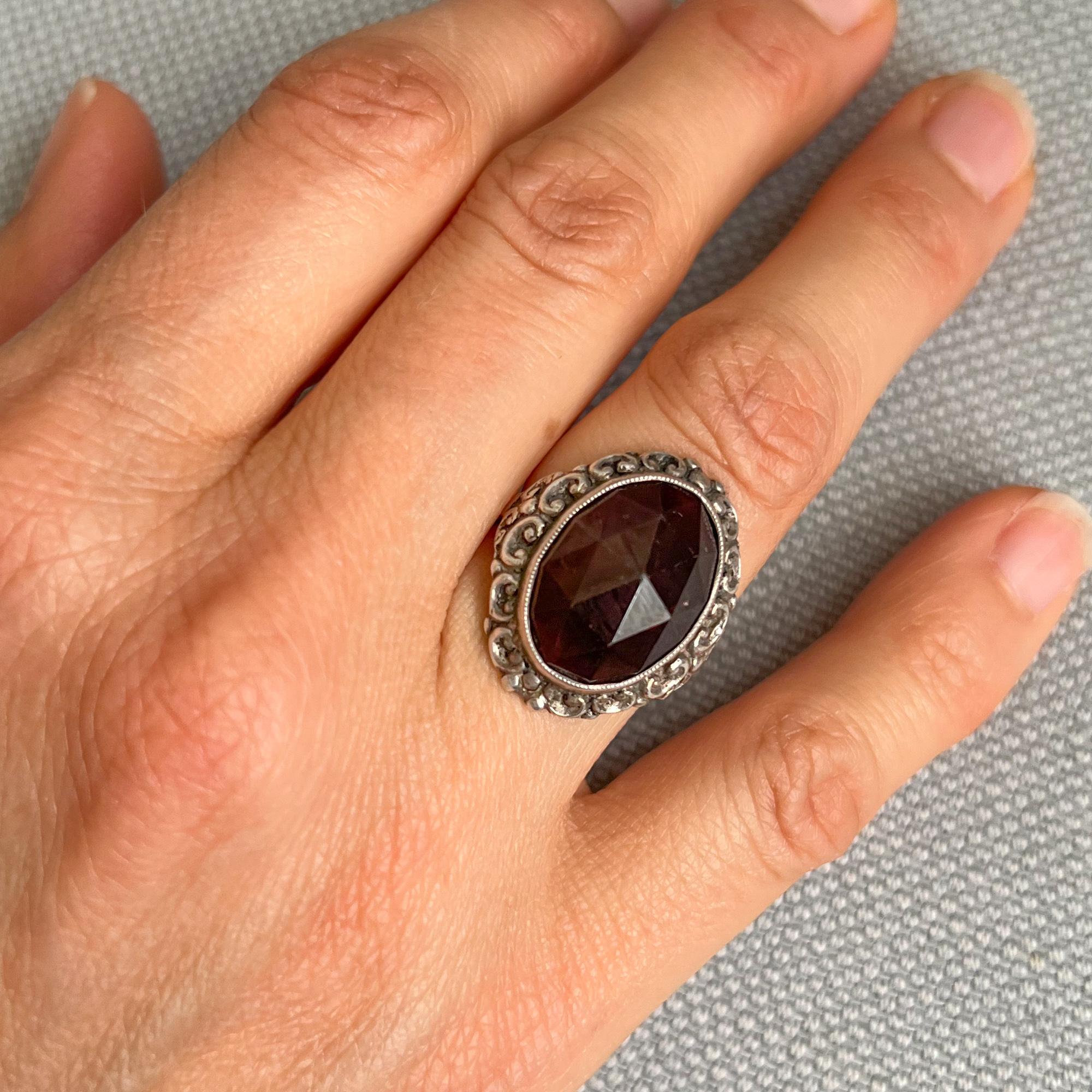 A preloved garnet ring made in silver. The silver oval shaped ring is set with a faceted garnet stone. The ring has a great design with one large oval garnet stone set in a silver ornate border of chased and curled motifs. The beauty of gemstones,