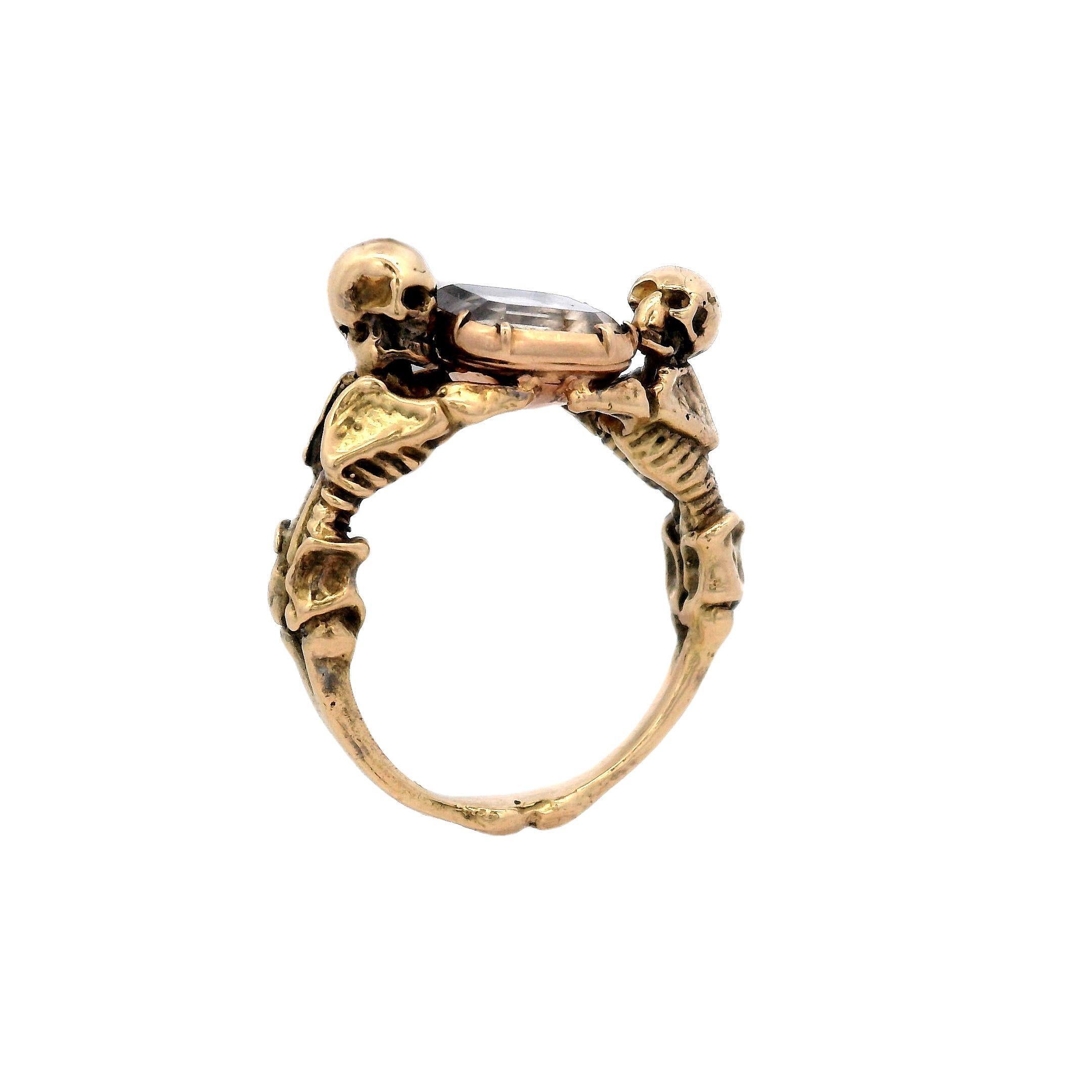 This Georgian style ring features two skeletons looking above and below while embracing a stuart crystal and is made from 18 karat gold.
This statement ring features a colorless rock crystal, elevated by two intricately detailed skeletons. The