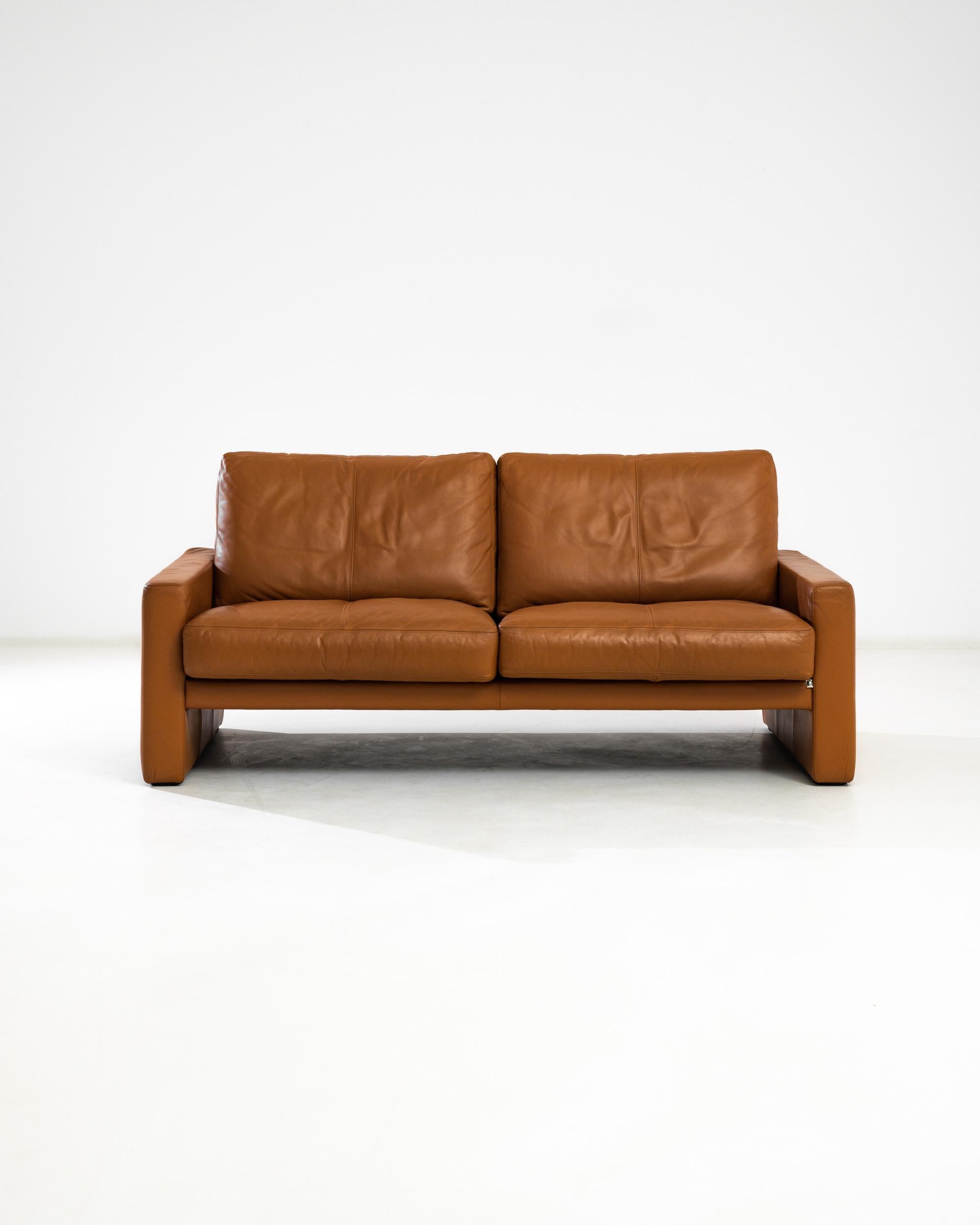 This two seat sofa was manufactured in the 20th century by the German furniture company WK Wohnen. Upholstered in high quality leather, it features comfortable plump seat cushions and smooth backrests supported by square arm panels on both sides.