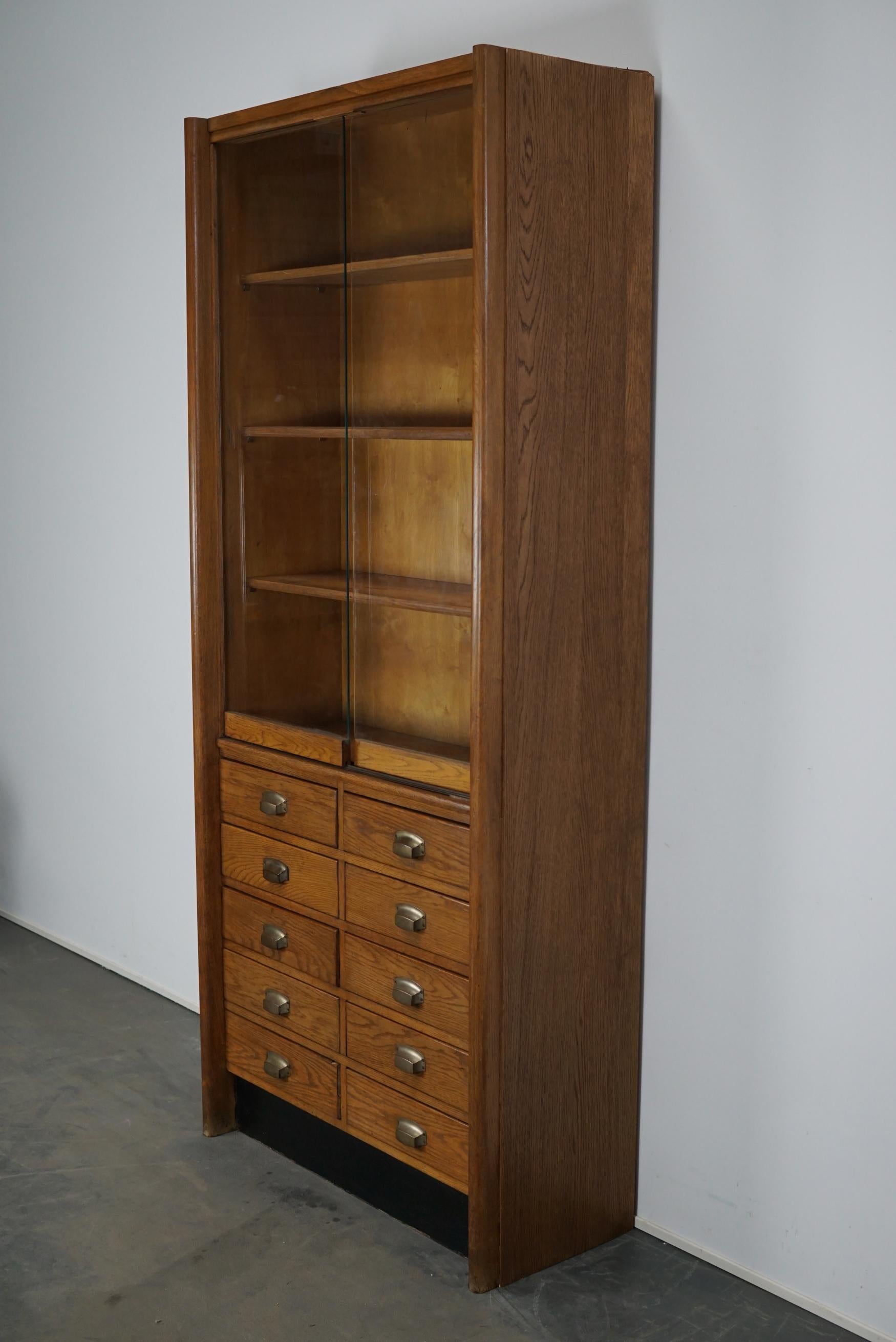 This apothecary cabinet was produced during the 1950s in Germany. This piece features 10 drawers with nice metal cup handles and two glass sliding doors with three shelves behind them. The interior dimensions of the drawers are: D x W x H 29 x 39 x