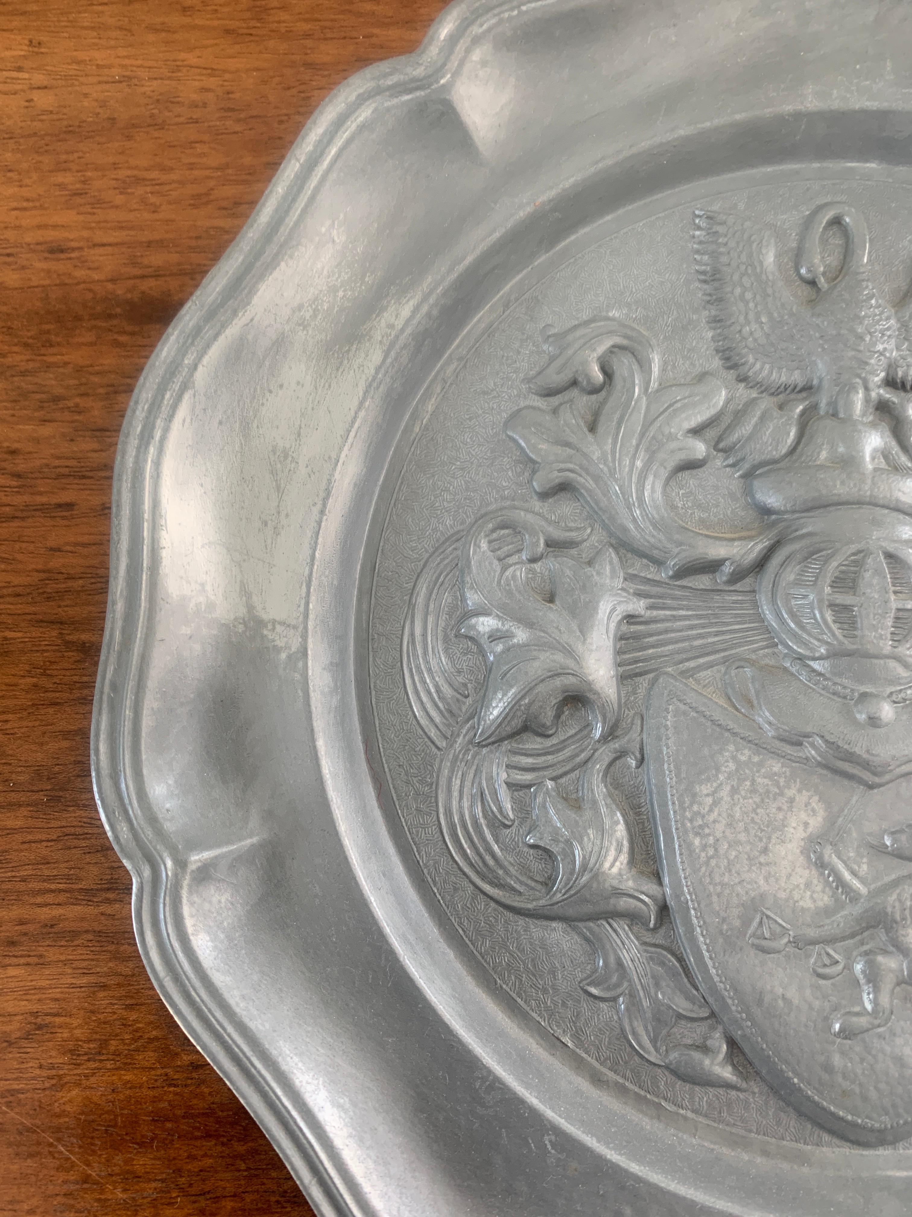 An early 20th century German pewter wall plate with a family crest consisting of a lion with sword on a shield culminating in a mother pelican feeding her babies. The symbolism of the mother pelican feeding her baby pelicans is part of an ancient