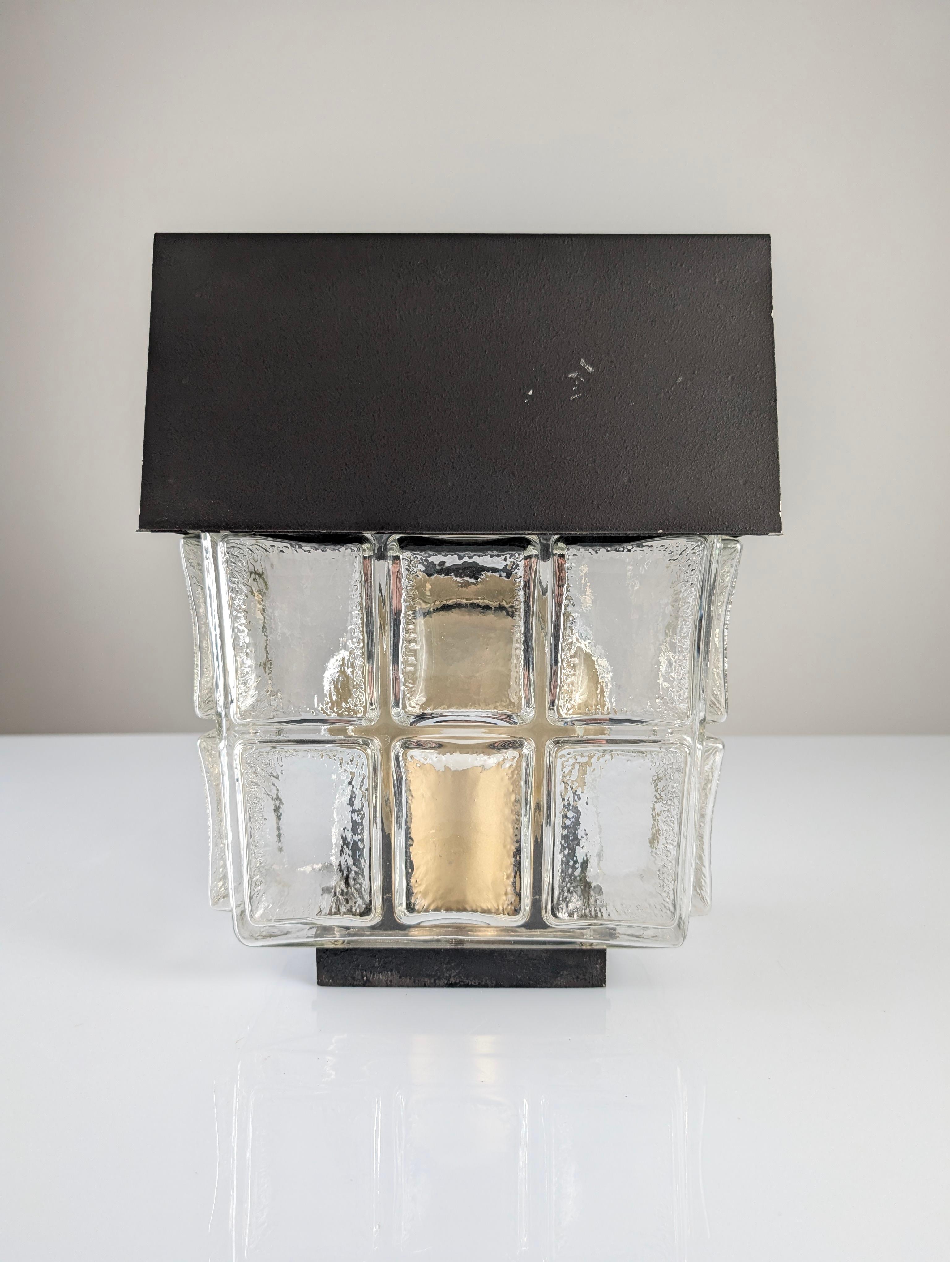 Vintage-Wandleuchte der deutschen Firma Bega aus den 1970er Jahren. Diese Wandleuchte für den Innen- und Außenbereich hat ein geradliniges Design mit geprägtem Glas und einer schwarzen Metallplatte. Auf der Rückseite befindet sich ein Kreis, der als