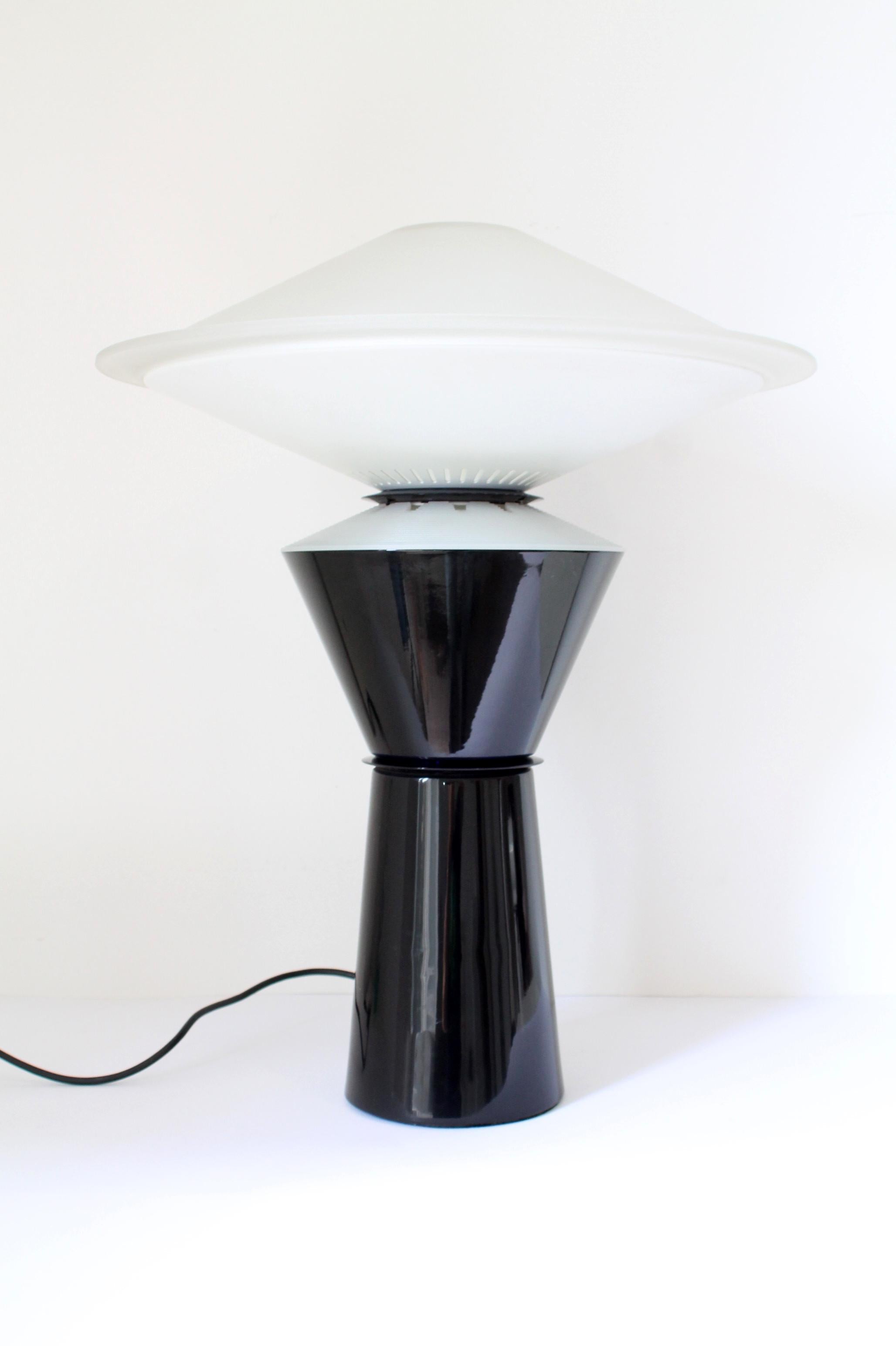 1980s Arteluce “Giada” table lamp by Pier Giuseppe Ramella.
A black lacquered metal base with a beautiful inner blue/purple lighted rim supports two thick frosted glass shells.

The switch allows enlighting separately the base and the lower side