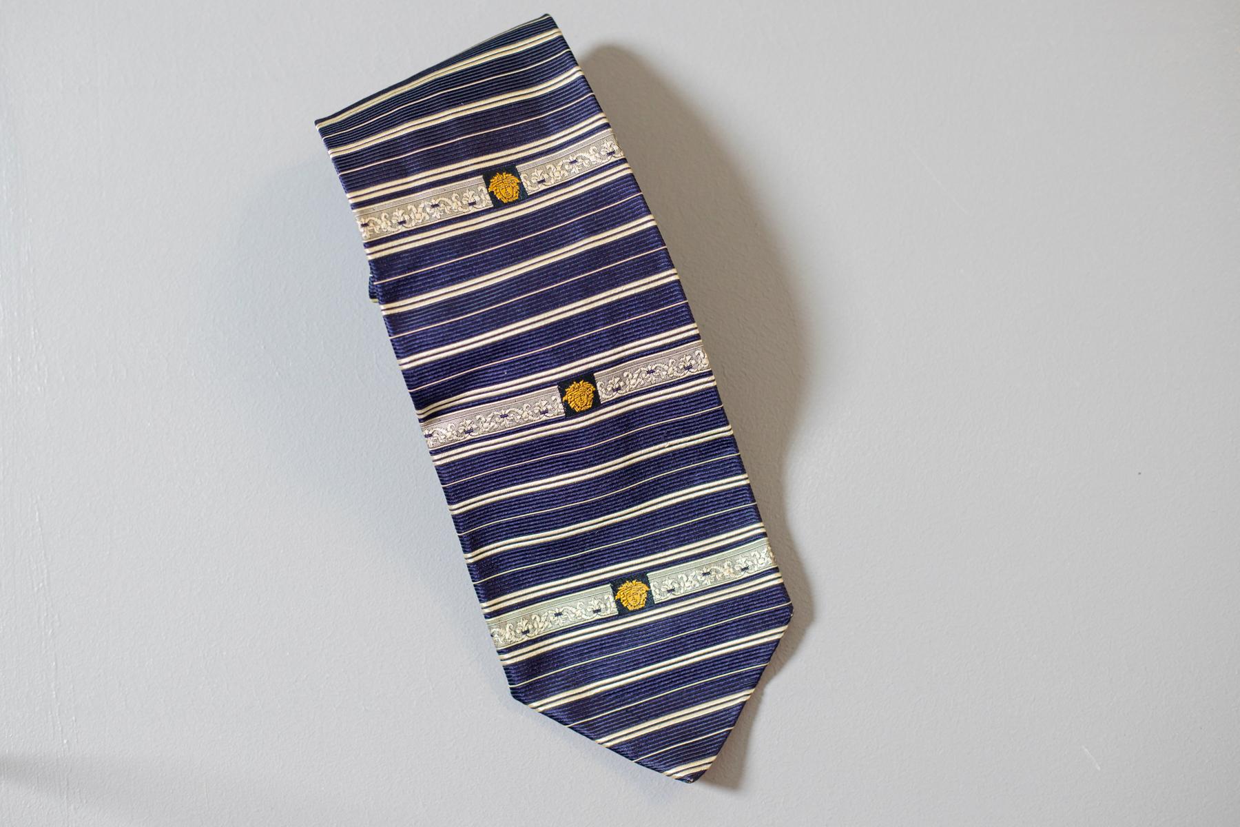 Cette cravate vintage conçue par Gianni Versace est un classique. Cette élégante cravate tout en soie est décorée non seulement de rayures bleues et blanches, mais aussi du symbole principal de Versace : La tête dorée de Méduse. Cette pièce de mode