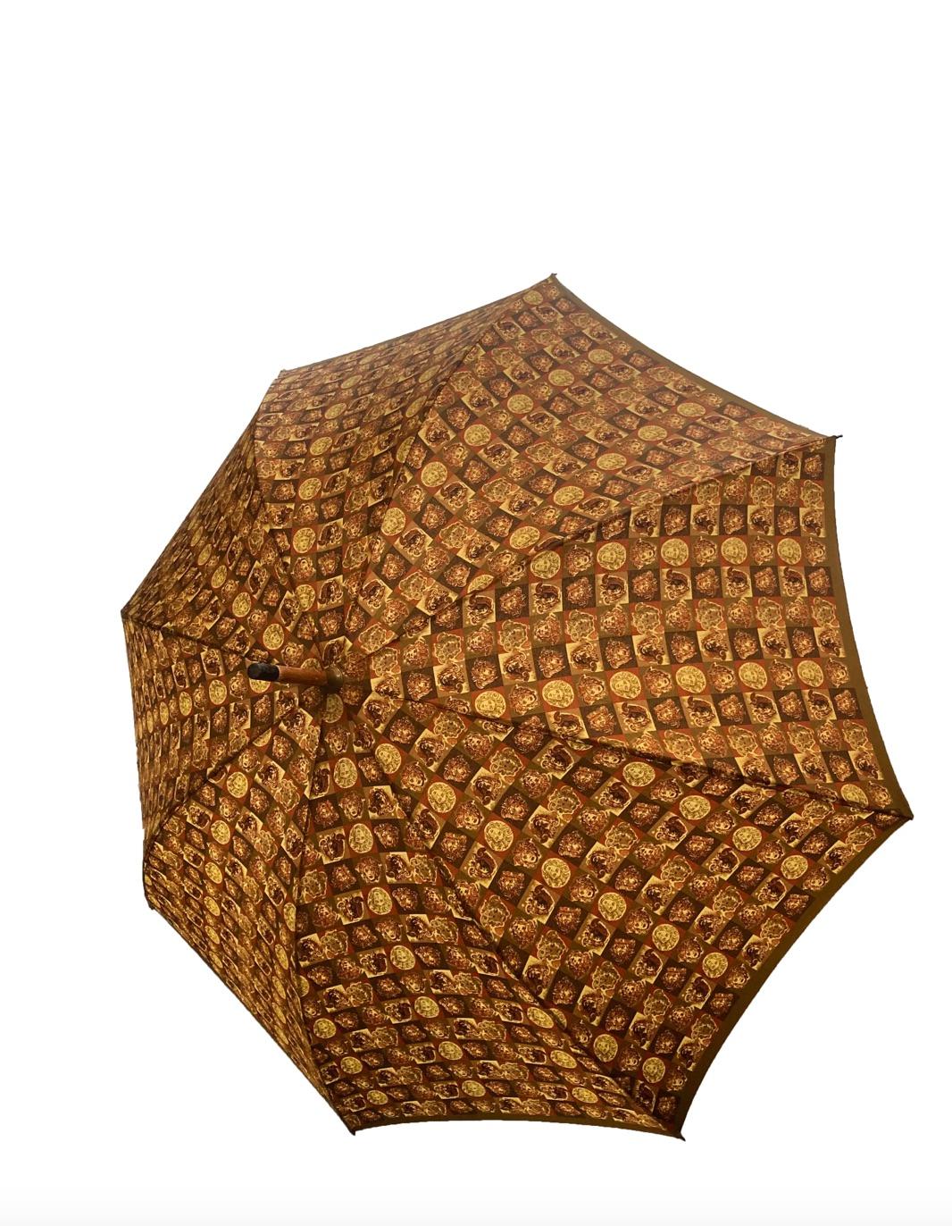 Parapluie vintage Gianni Versace de la marque éponyme. Manche en bois brun doré avec poignée en bois crochetée, ornée du logo de la tête de méduse découpée, entourée d'une bordure de style grec en métal doré. La tige en nylon kaki arbore un motif de
