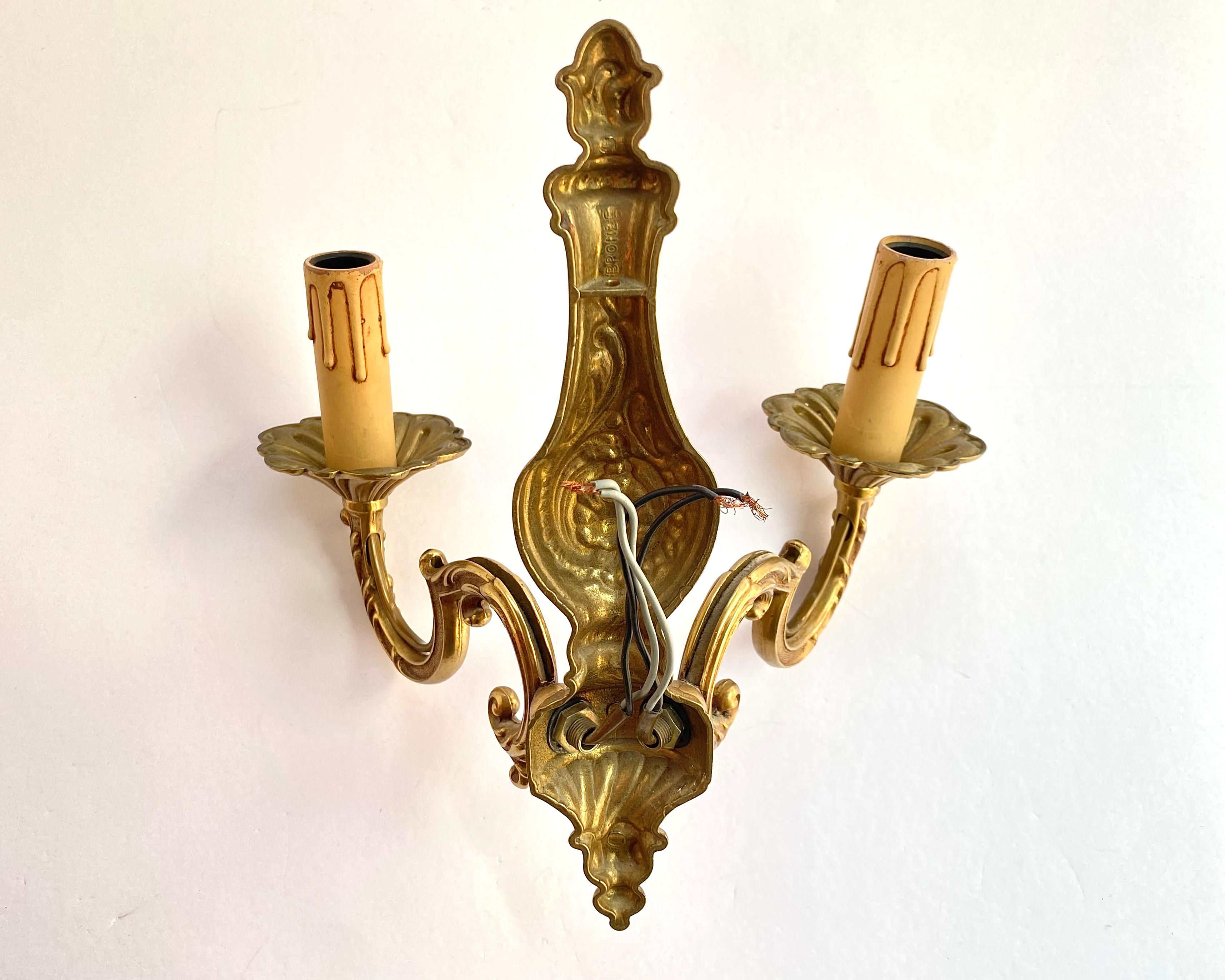 Lampe murale Vintage Twin-Arm en bronze de France imitant les candélabres classiques, circa 1960.

Il y a une marque sur l'applique.u2028u2028

Le design du luminaire contient des éléments du style baroque. 

Les détails décoratifs en bronze moulé