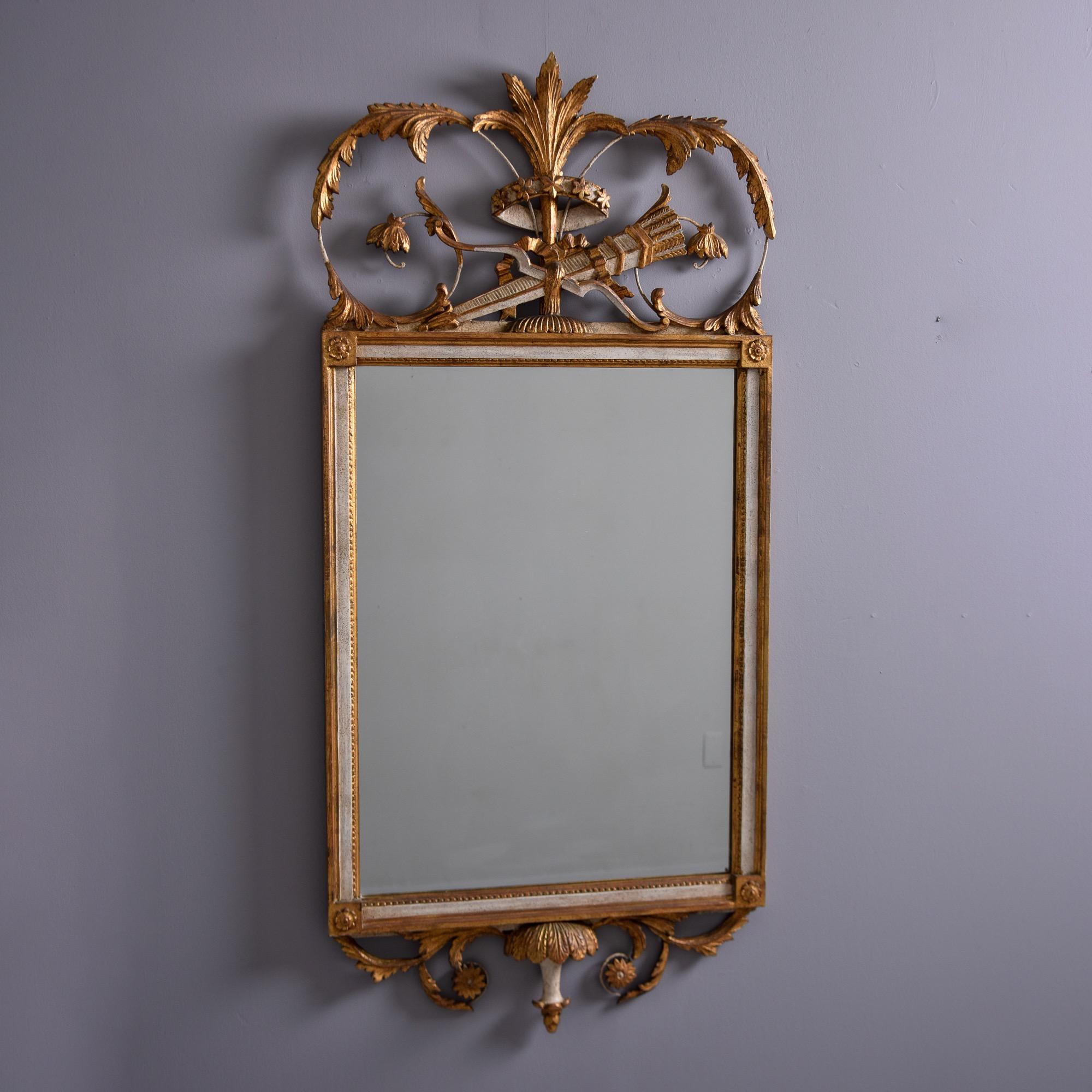 Found in the US, this circa 1950s decorative mirror features a gilt and cream wooden frame with an ornate crest. Rectangular mirror features a medallion with floral motif on each corner and a tall, open work crest with acanthus leaves and a torch.