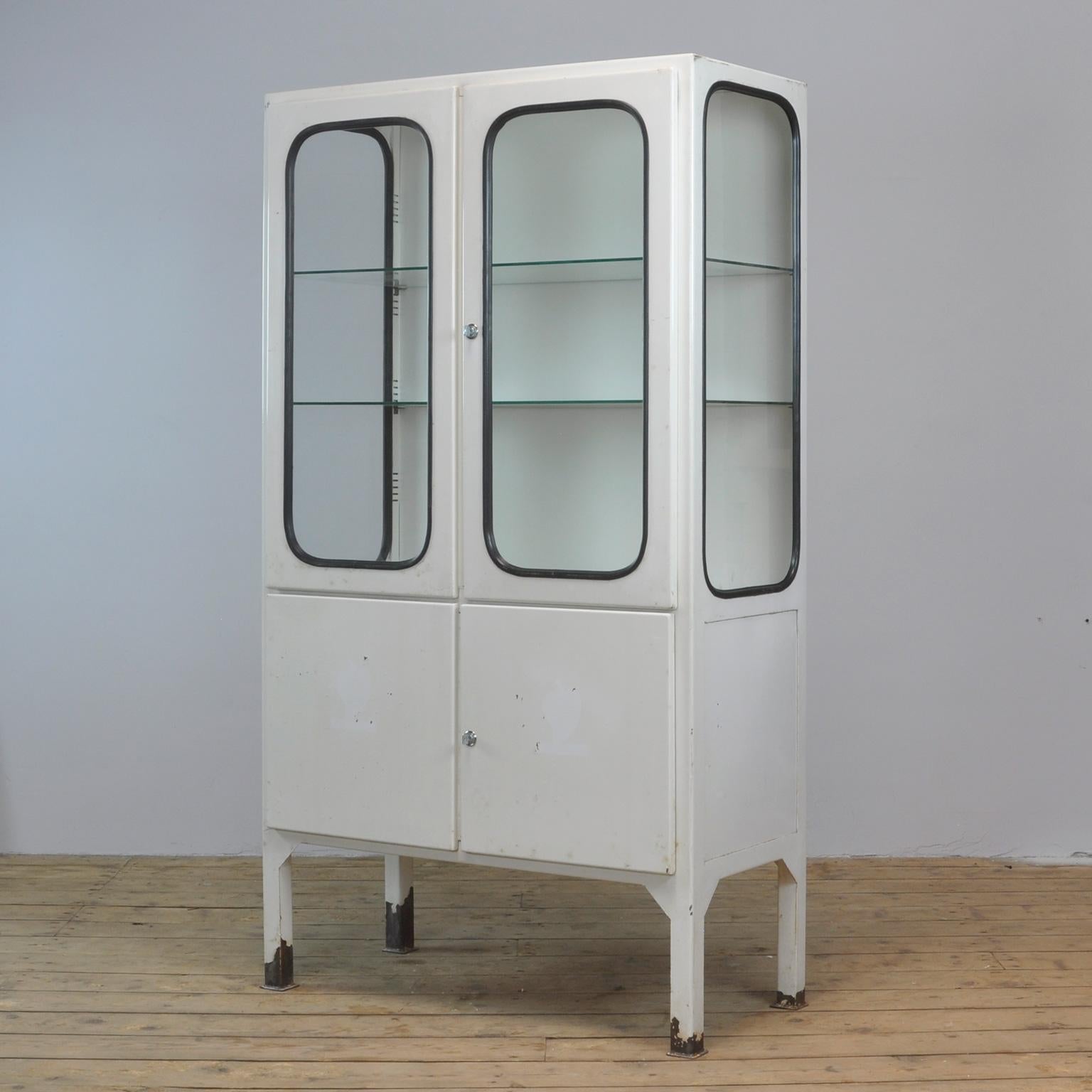 This medical cabinet was designed in the 1970s and was produced circa 1975 in Hungary. It is made from iron and glass, and the glass is held by a black rubber strip. The cabinet features two adjustable glass shelves and functioning locks. The