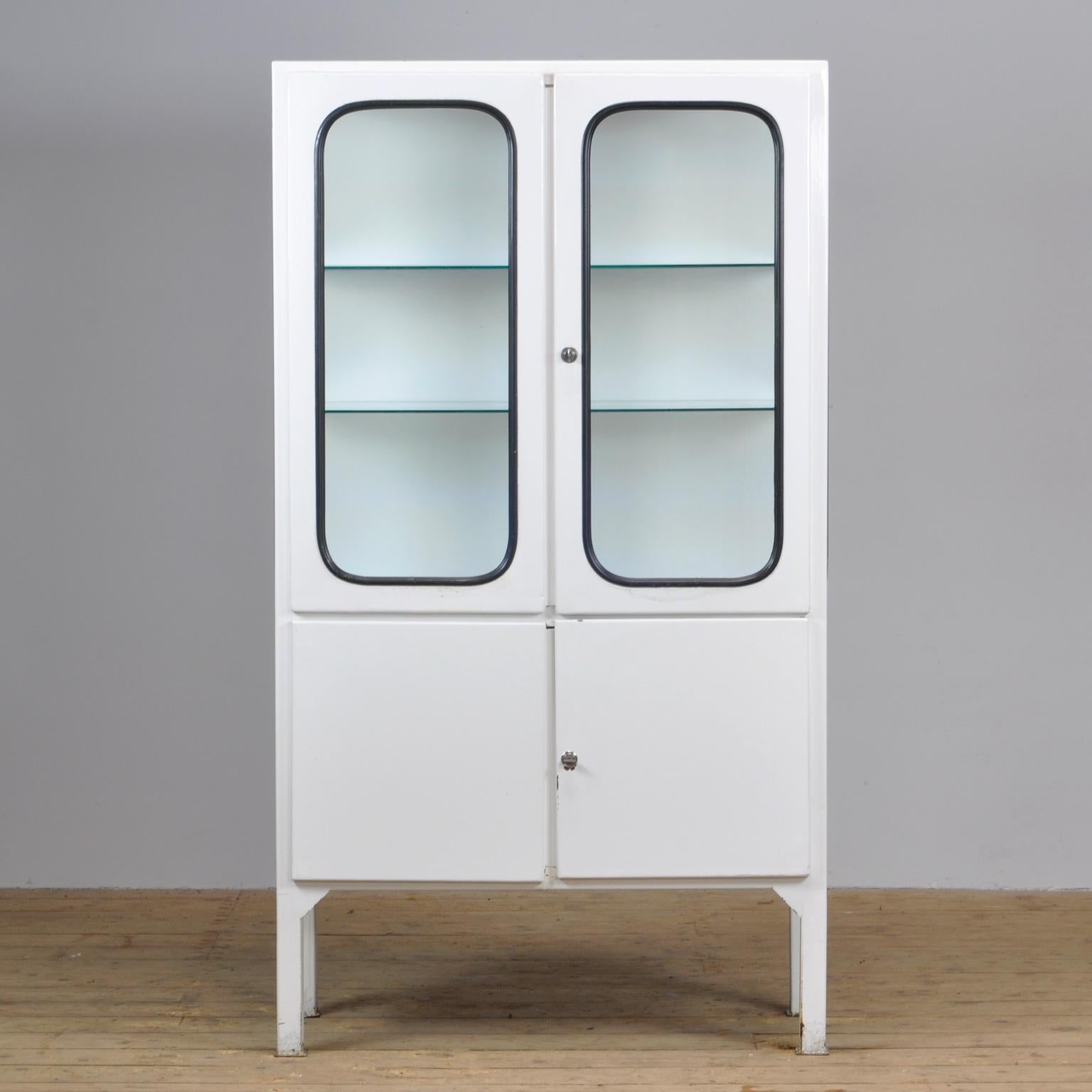 This medical cabinet was designed in the 1970s and was produced circa 1975 in Hungary. It is made from iron and glass, and the glass is held by a black rubber strip. The cabinet features two adjustable glass shelves and functioning locks.