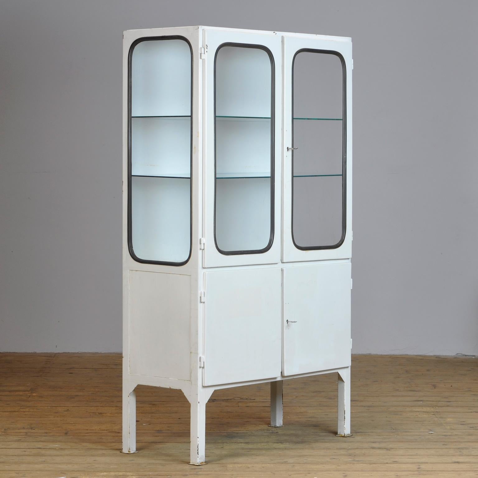 This medical cabinet was designed in the 1970s and was produced circa 1975 in Hungary. It is made from iron and glass, and the glass is held by a black rubber strip. The cabinet features two adjustable glass shelves and functioning locks.
  