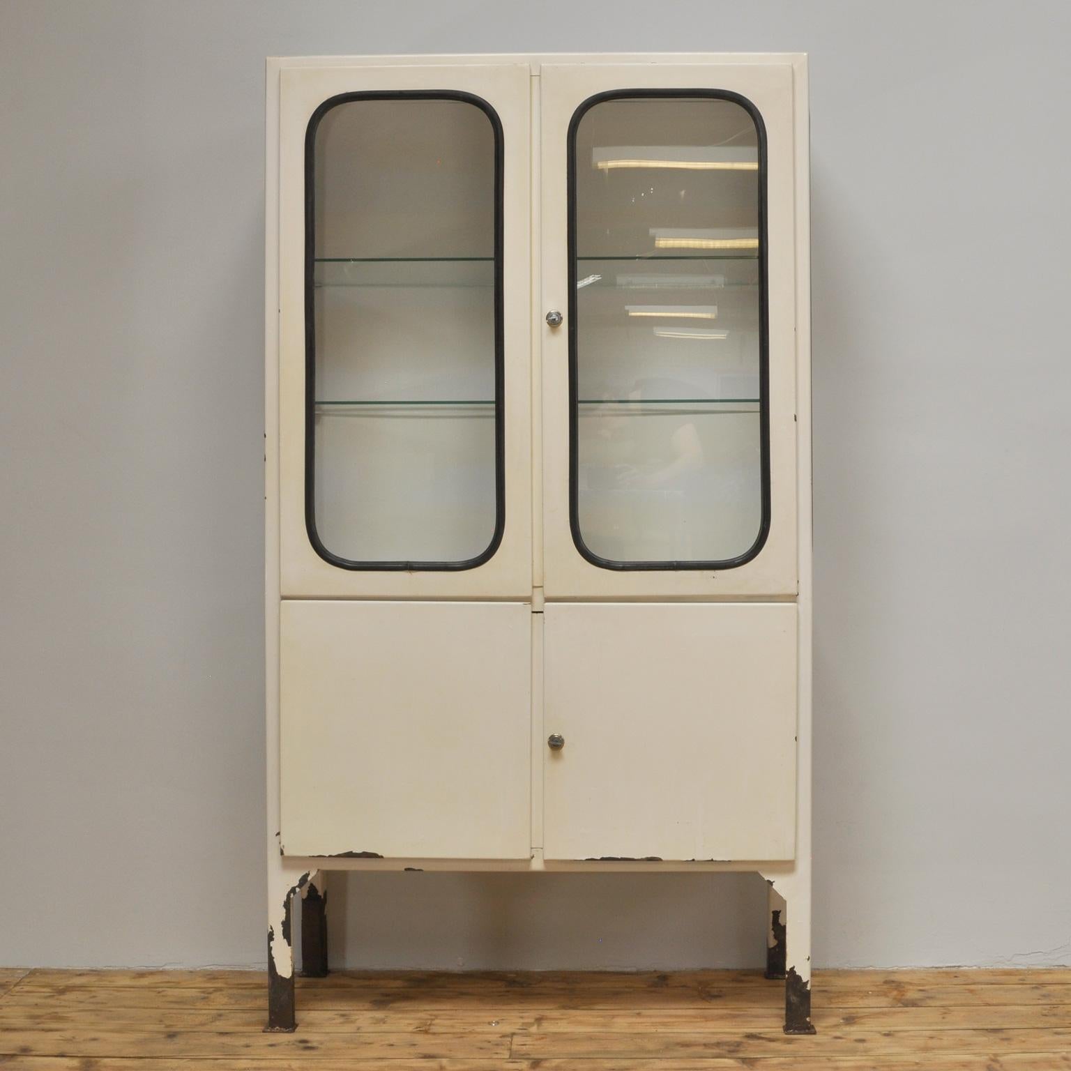 This medicine cabinet was designed in the 1970s and was produced circa 1975 in Hungary. It is made from iron and glass, and the glass is held by a black rubber strip. The cabinet features two adjustable glass shelves and functioning locks.