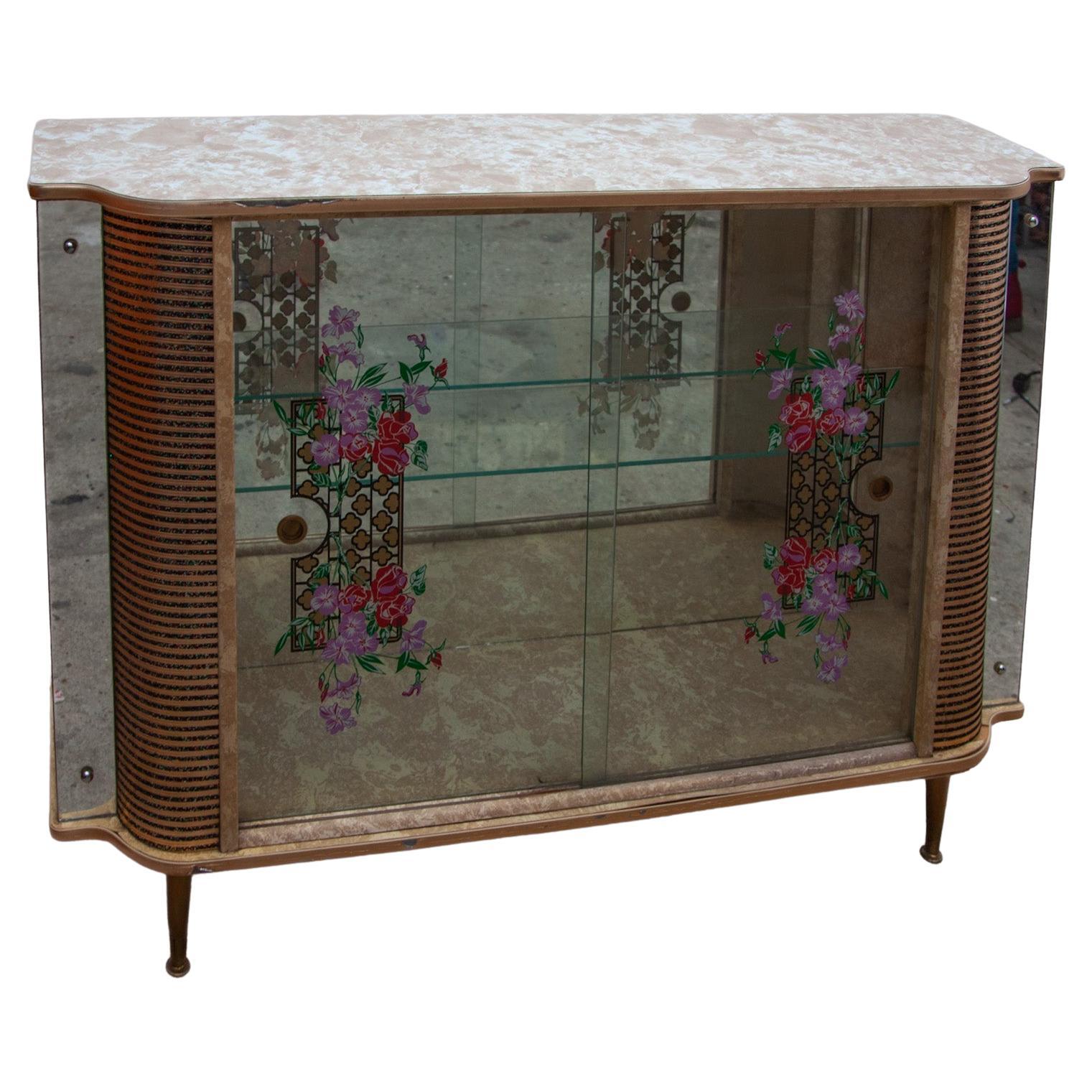 A vintage glass cocktail display cabinet with glass doors and shelves. Decorative illustrated glass in a teak frame with two glass shelves and mirrored accents. Brass trim throughout. In good original condition this piece of furniture has certainly