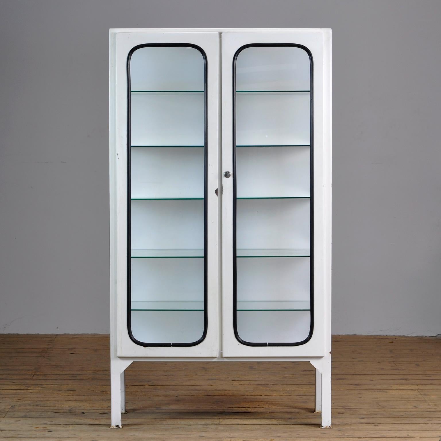 This medicine cabinet was designed in the 1970s and was produced circa 1975 in Hungary. It is made from iron and glass, and the glass is held by a black rubber strip. The cabinet features five adjustable glass shelves and a functioning lock.