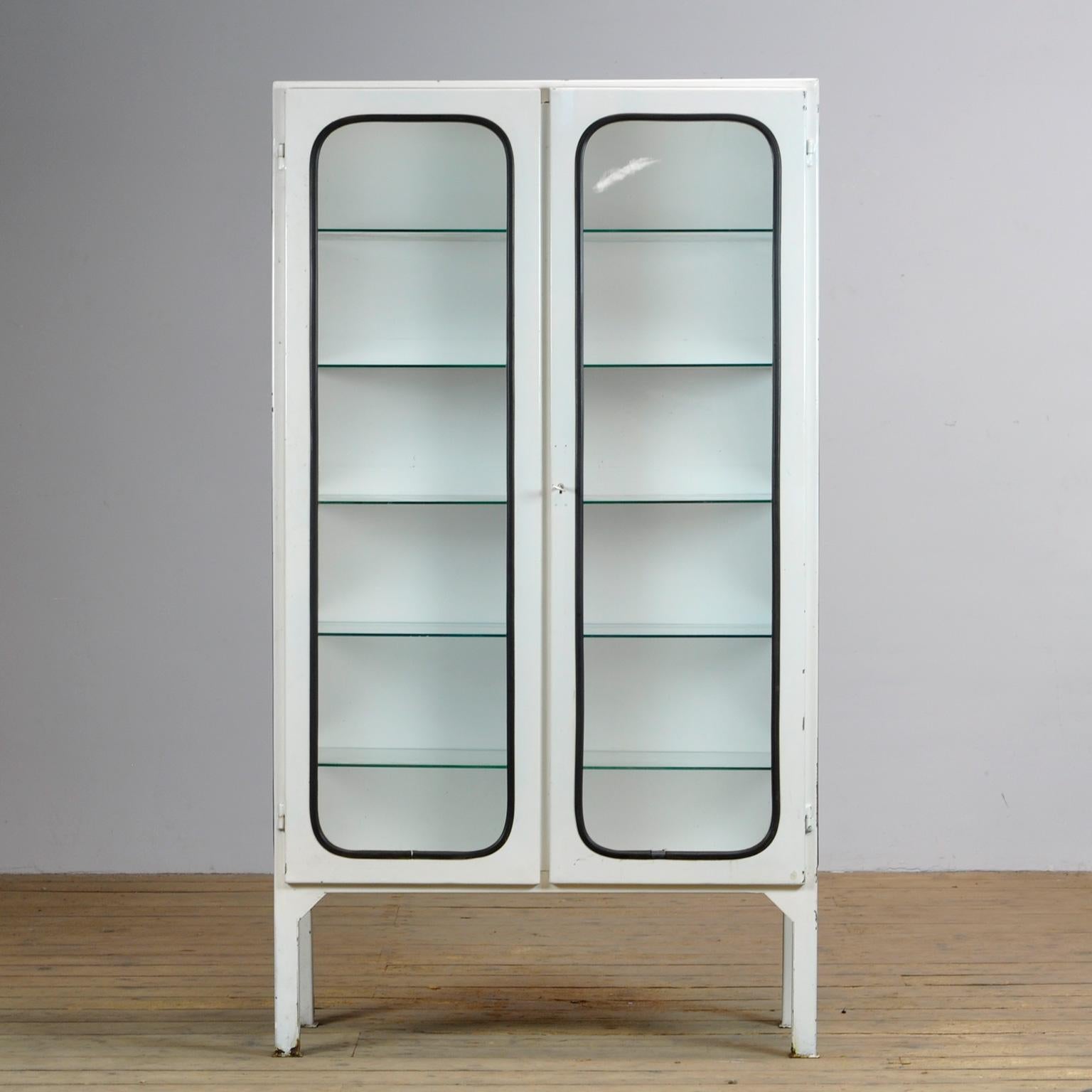 This medicine cabinet was designed in the 1970s and was produced circa 1975 in hungary. It is made from iron and glass, and the glass is held by a black rubber strip. The cabinet features five adjustable glass shelves and a functioning lock.