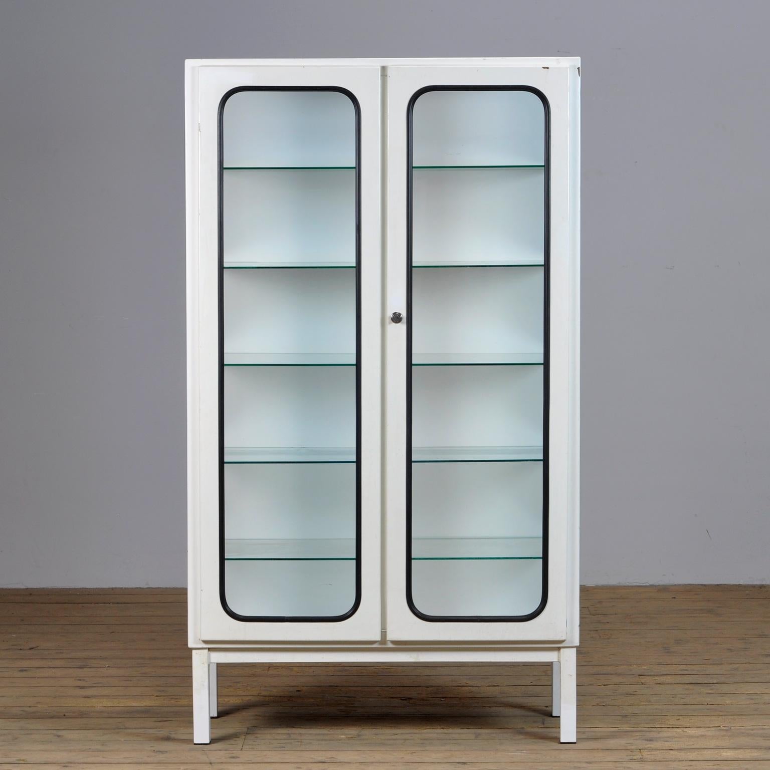 This medicine cabinet was designed in the 1970s and was produced circa 1975 in hungary. It is made from iron and glass, and the glass is held by a black rubber strip. The cabinet features five adjustable glass shelves and a functioning lock.
