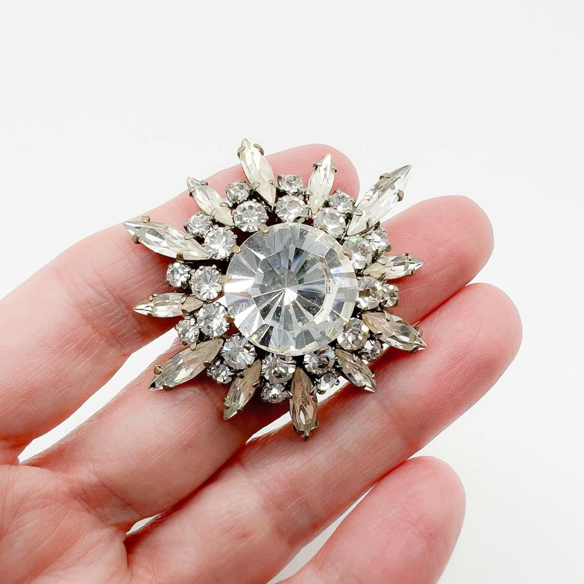 A Vintage Glass Paste Starburst Brooch. Crafted around the 1930s this delightful pin features a gigantic solitaire crystal surrounded by an array of chaton and marquise crystals conjuring the illusion of starburst. The glint and glisten factor is