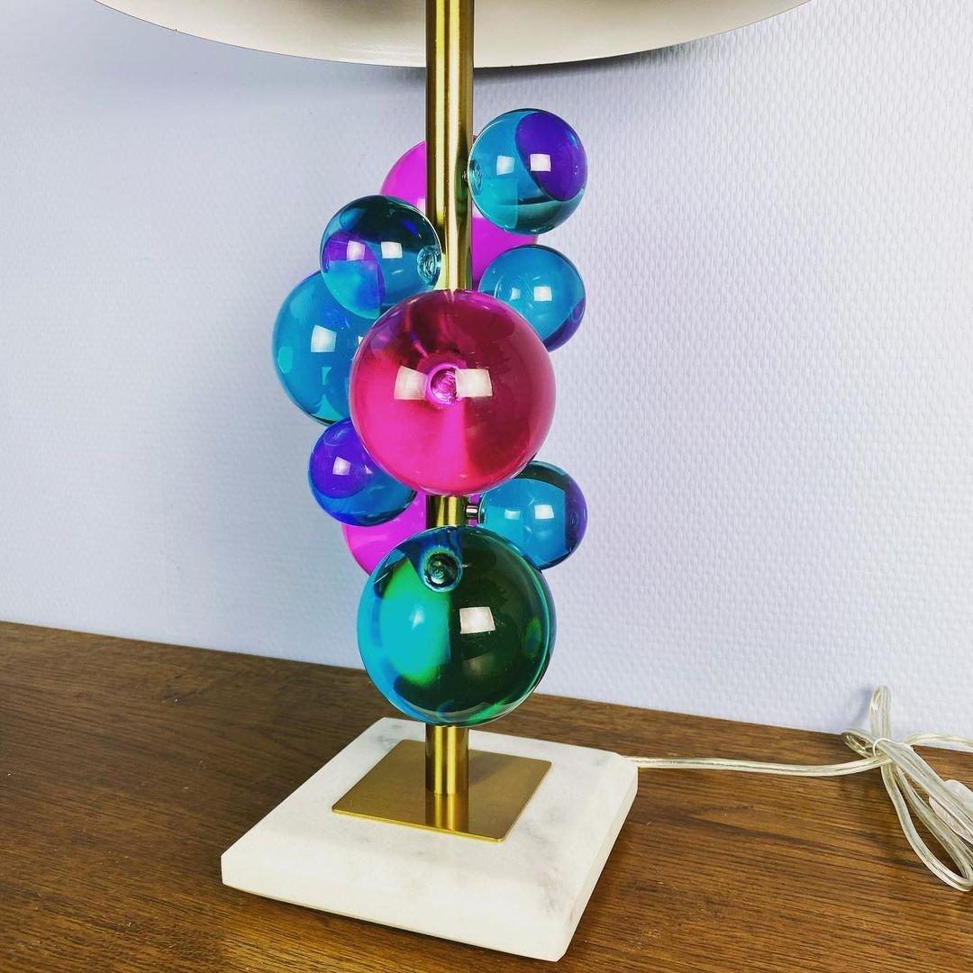 - The Globo table lamp by Jonathan Adler is a constellation of multicolored acrylic spheres that float on a thin brass stem fixed to a solid marble base and topped with a brass dome.
- Circa 2010s.