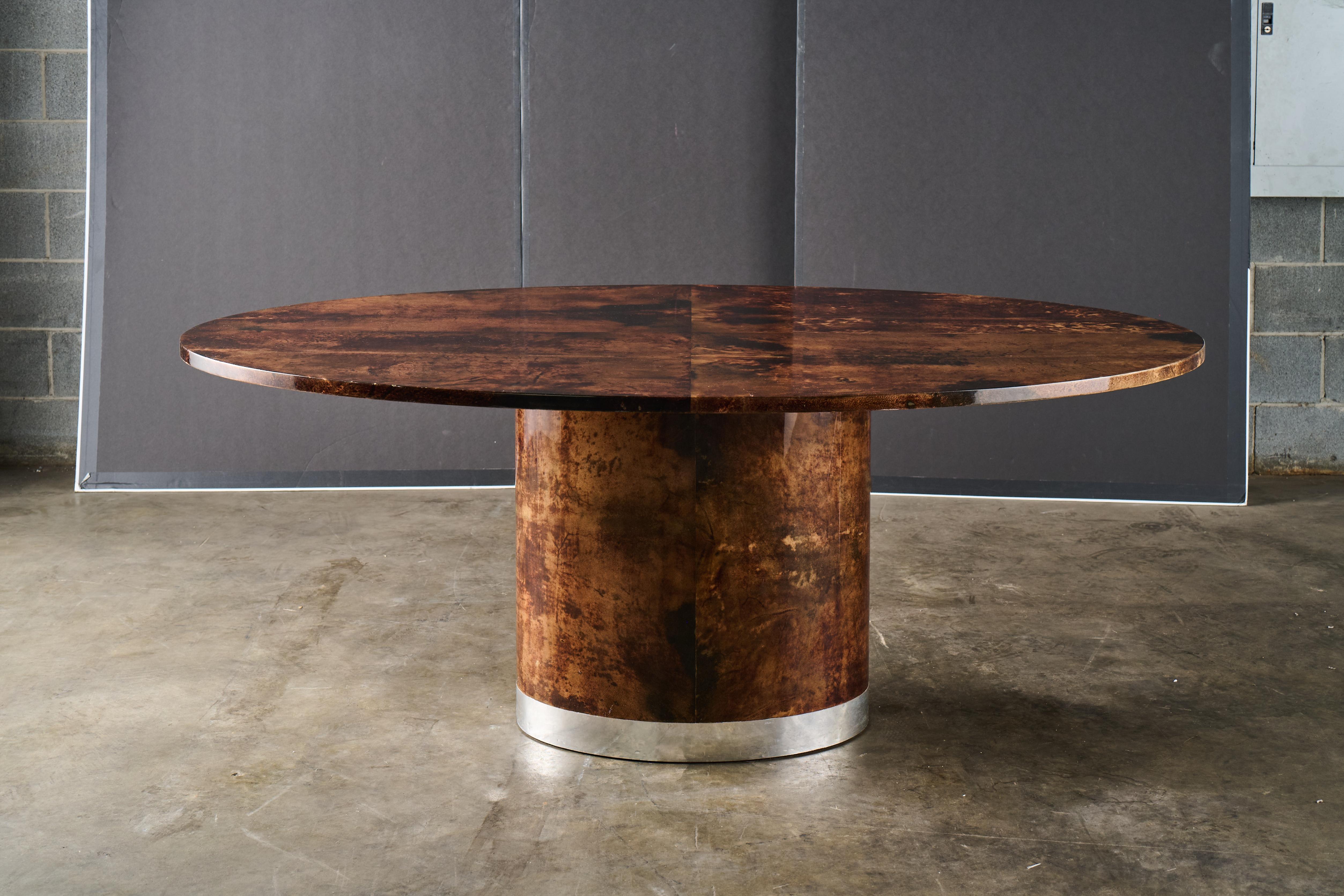 This oval table features rich goatskin with a lacquer finish, giving it a luxurious, almost liquid depth and pattern. Its cylindrical pedestal base provides a sculptural presence and is finished with a brushed chrome or stainless steel band along