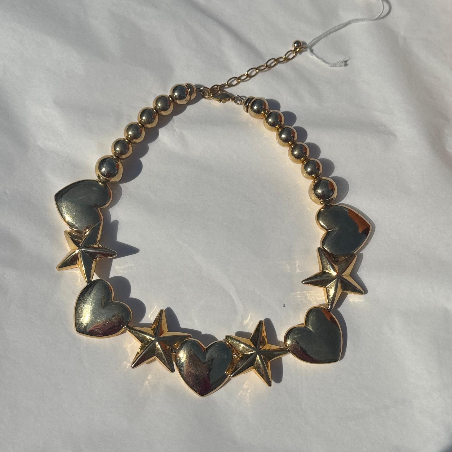 Bold statement necklace in shiny gold-tone metal, featuring an alternating pattern of puffed heart and star-shaped elements that create a playful and eye-catching rhythm. The bright, sculptural design is typical of 1980s and 1990s costume jewelry-an