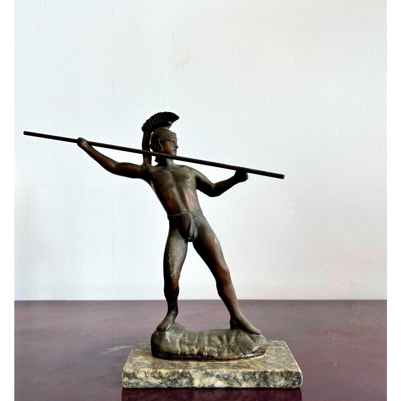 Questa figura in bronzo cattura l'intensità di un guerriero classico, fermo nel momento prima di lanciare la sua lancia. Indossando solo un elmo corinzio e un perizoma, la sua forma snella e la sua posizione equilibrata evocano l'atletismo e