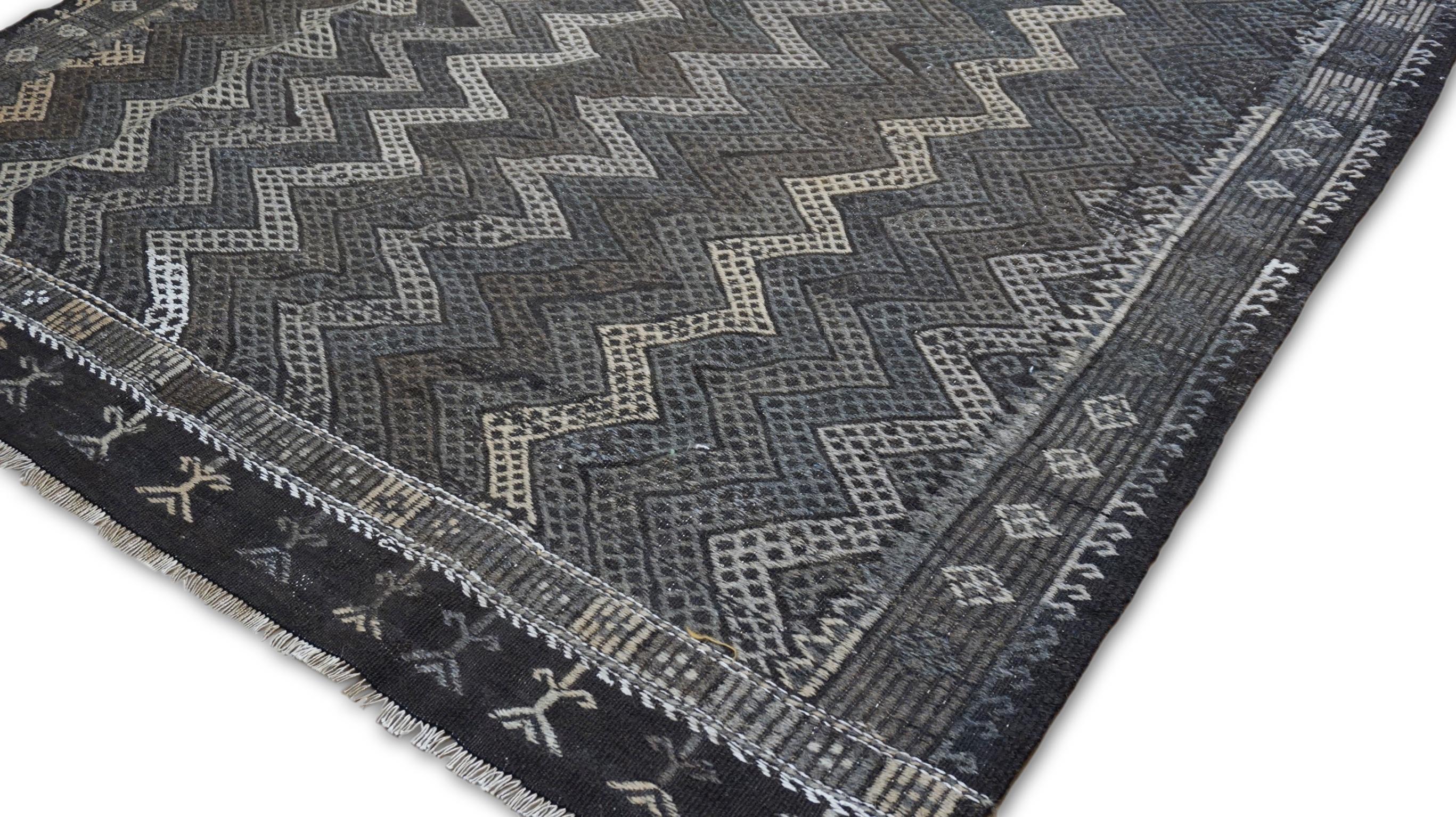 Questo affascinante tappeto kilim vintage in lana presenta un intricato motivo tribale a zig zag, magistralmente intrecciato nelle tonalità del grigio, del marrone e dell'avorio. Il design geometrico è impreziosito da un dettagliato reticolo di