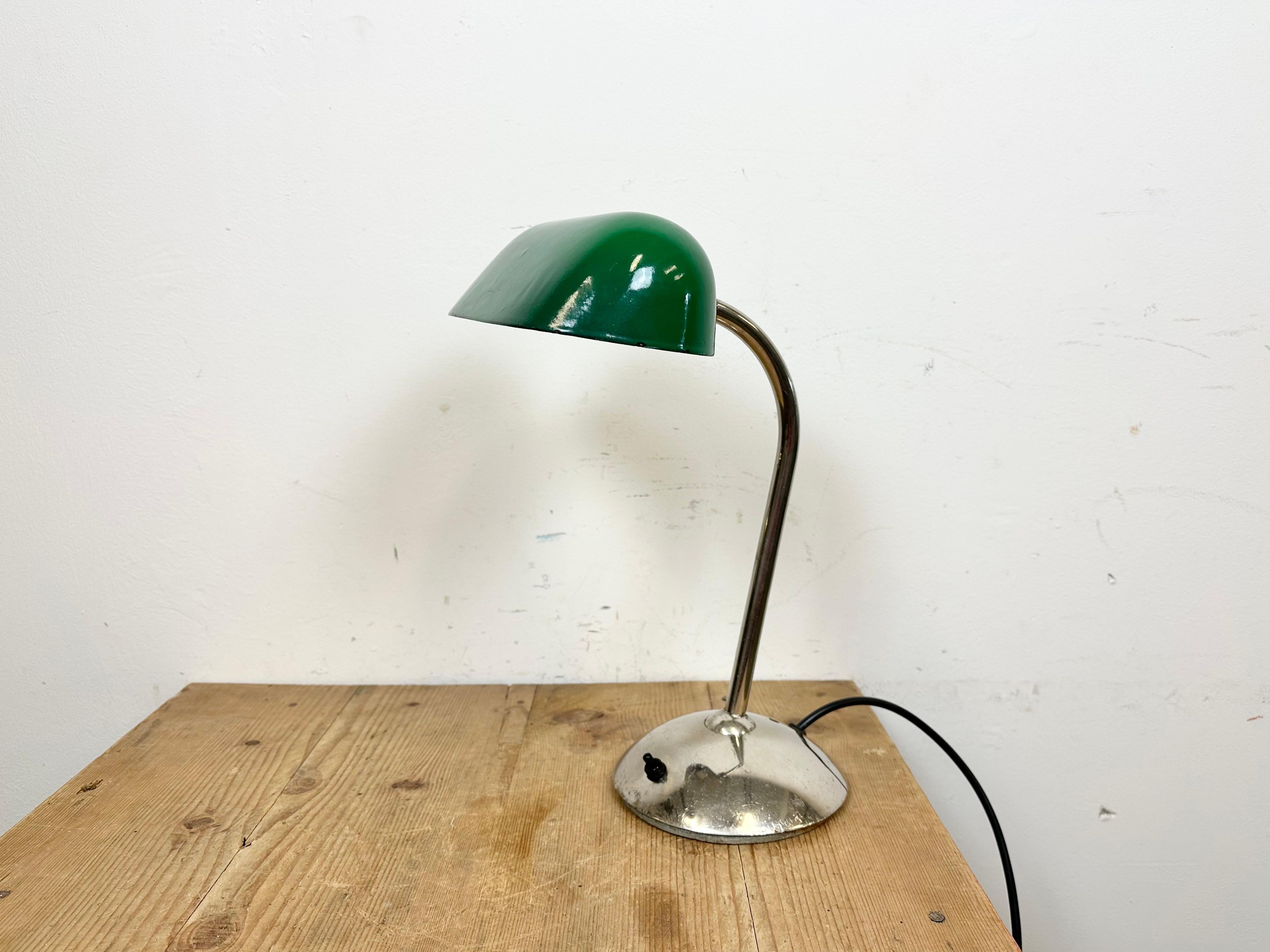 This green table lamp was made in former Czechoslovakia during the 1950s. It features a green enamel shade with a white enamel interior and a chrome plated iron base and arm with two adjustable joints. Original porcelain socket requires E 27/ E 26