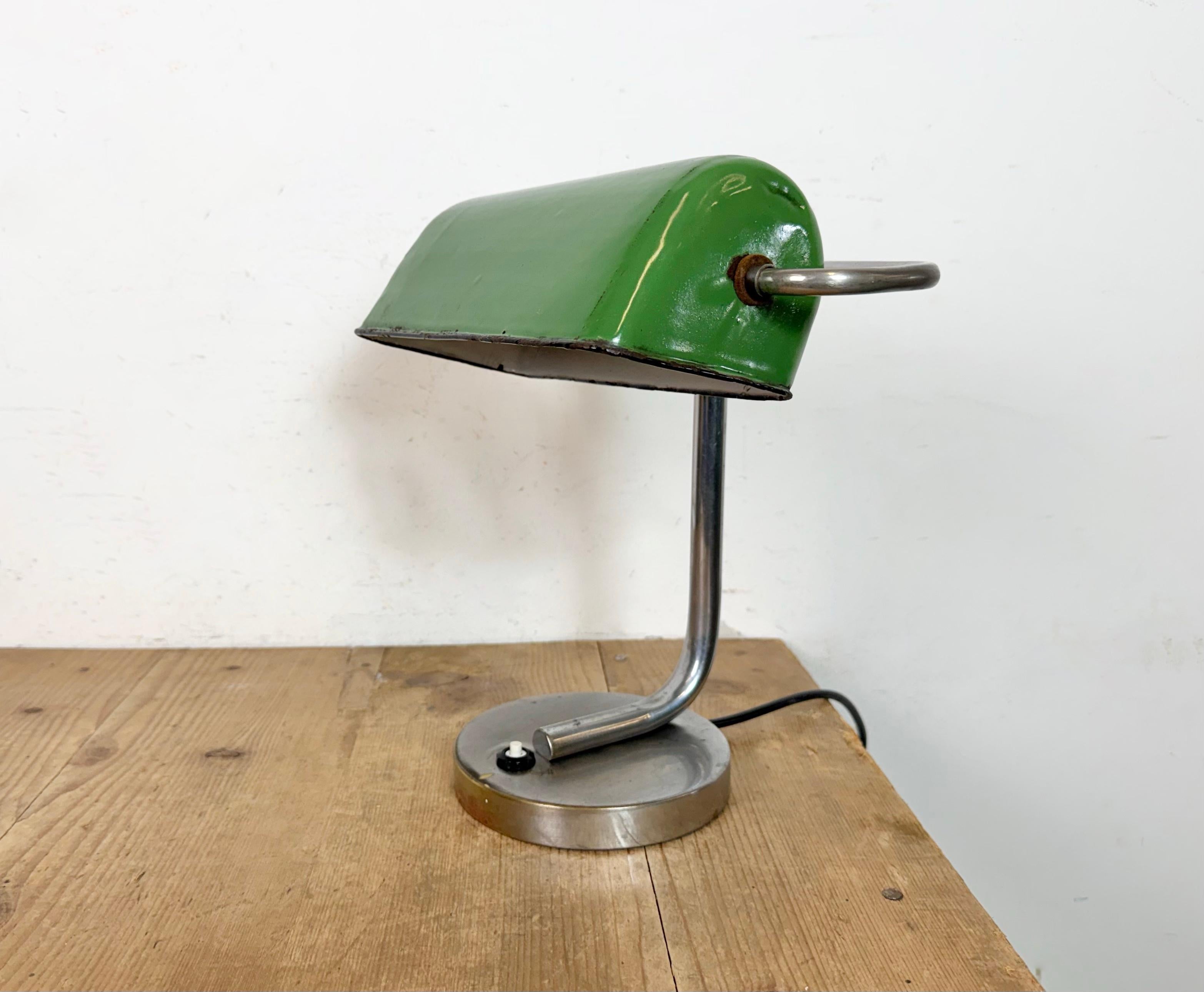 This vintage banker table lamp was made in former Czechoslovakia during the 1950s. It features a green enamel lampshade with a white enamel interior and a chrome plated iron arm and base with switch.. The original bakelite socket requires a standard