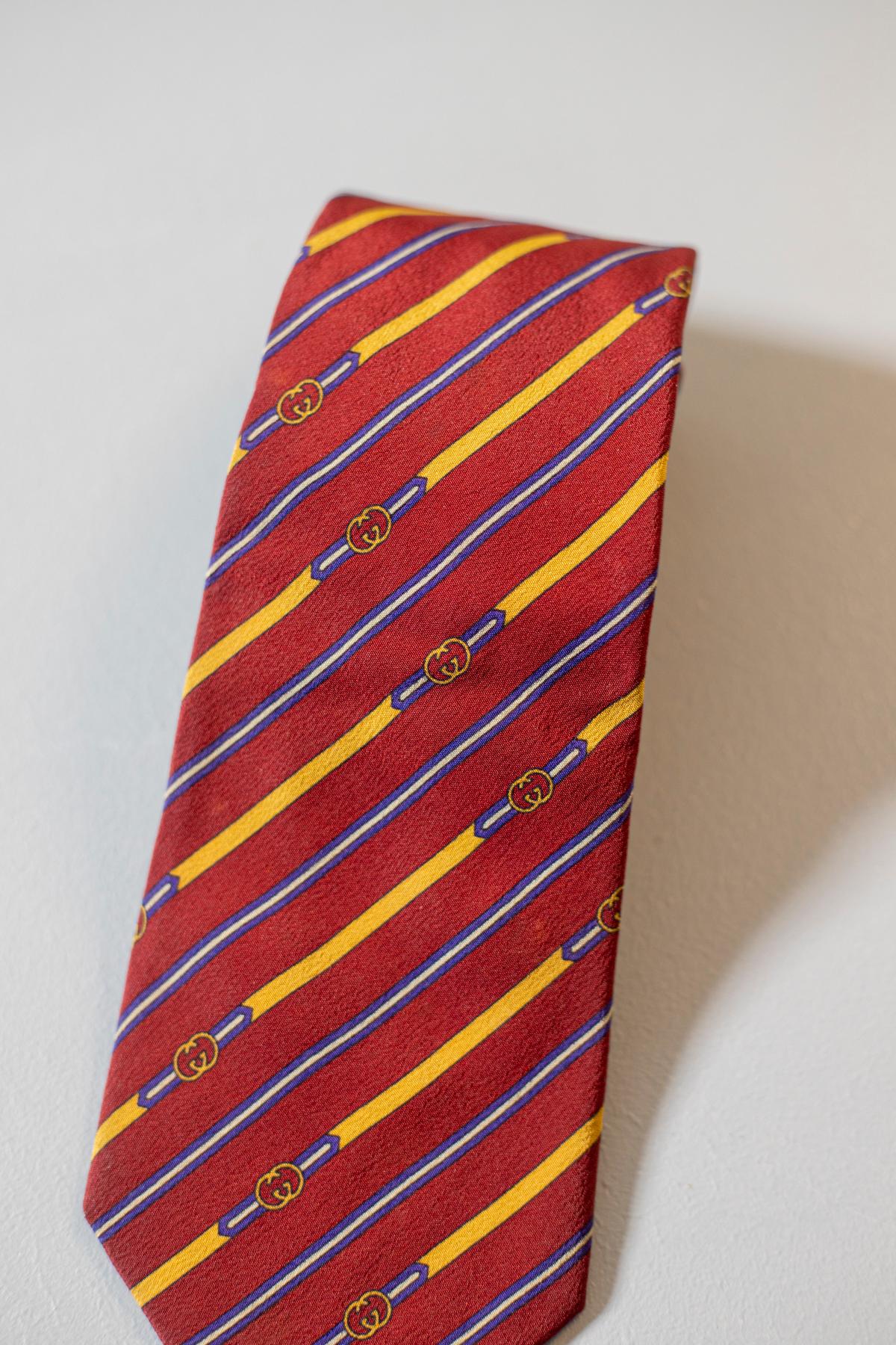 Diese Vintage-Krawatte aus Seide wurde von Gucci entworfen. Dieses Accessoire ist elegant und stilvoll. Es zeigt blaue, gelbe und weiße Streifen auf einem roten Hintergrund. Es ist perfekt für einen eleganten Anlass. 