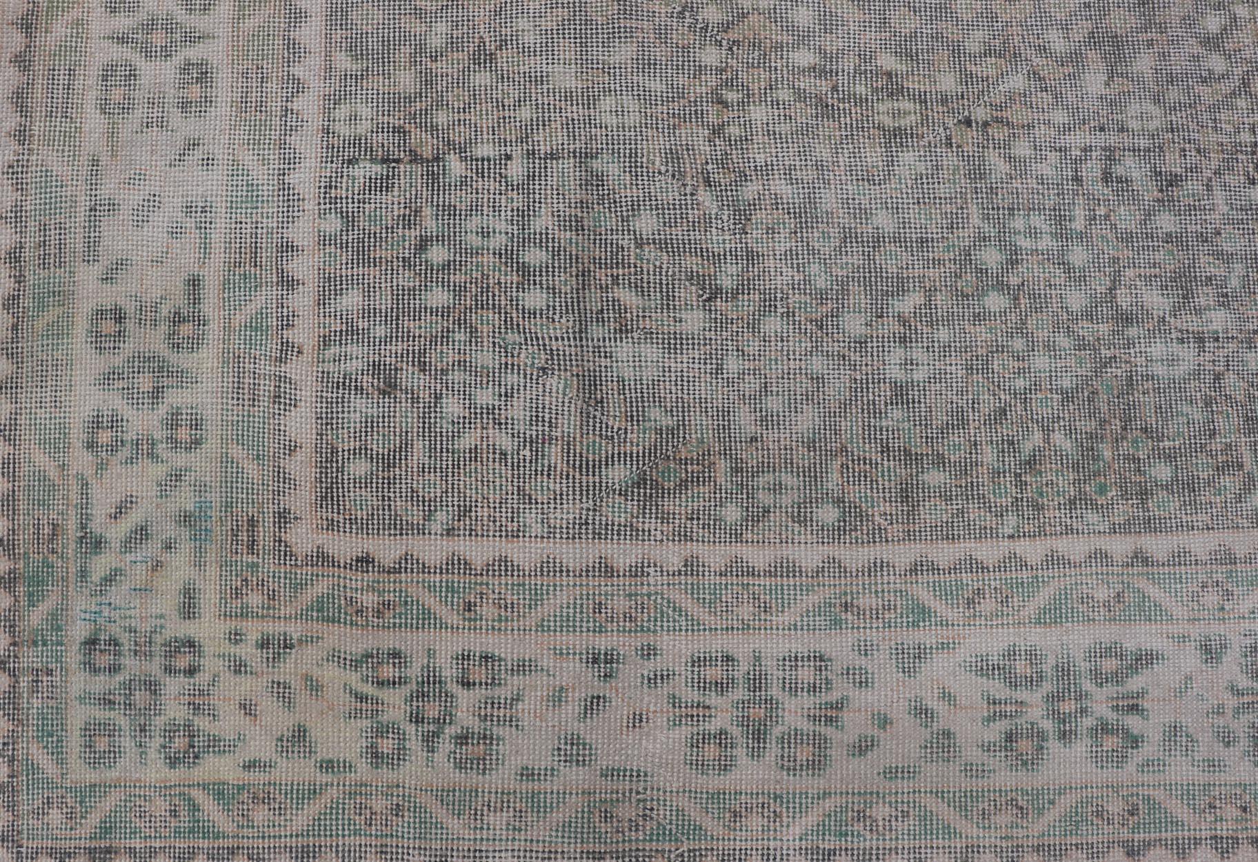 This vintage Turkish Oushak rug has been hand-knotted in wool and features an all-over sub-geometric distressed floral design rendered in cream, blue, green and ivory tones. A complementary, multi-tiered border encompasses the entirety of the piece;
