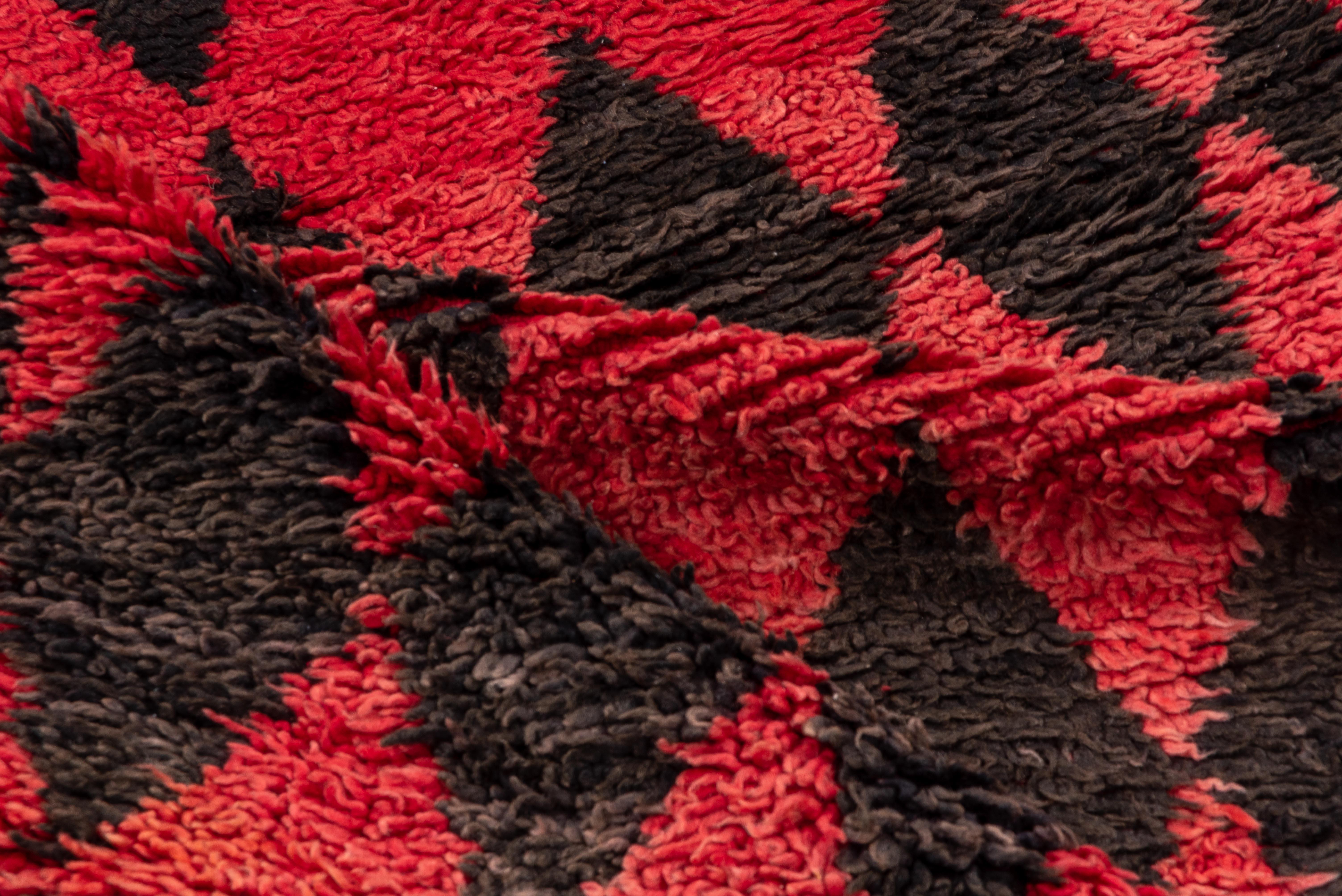 Este llamativo camino de mesa marroquí vintage presenta un atrevido motivo geométrico compuesto por rombos rojos y negros alternados, que evoca una estética rítmica y arlequinada. Anudada a mano con un grueso pelo de lana, la alfombra ofrece una
